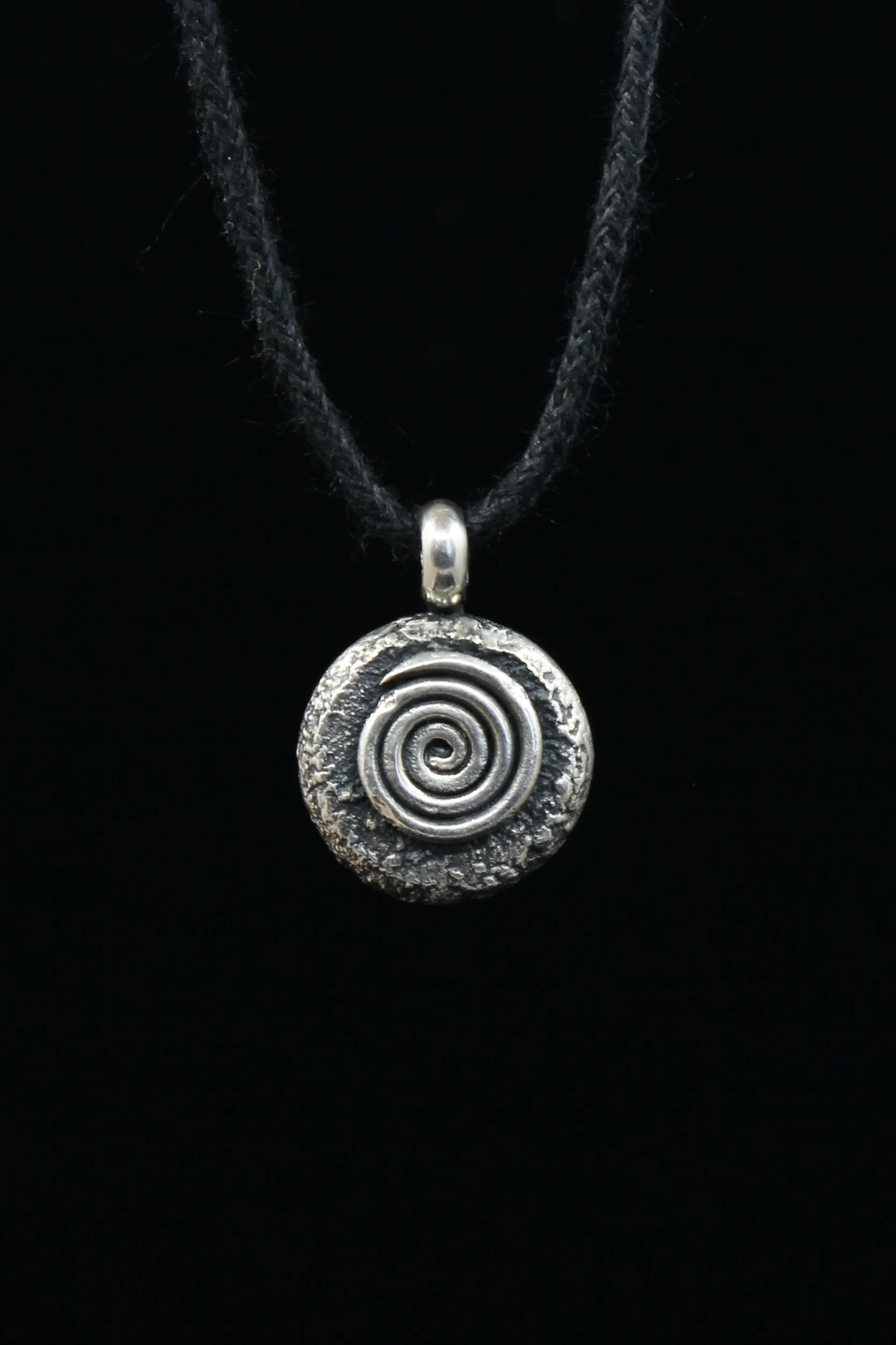 Hand-forged round silver pendant with raw oxidised texture and raised spiral design on black cord