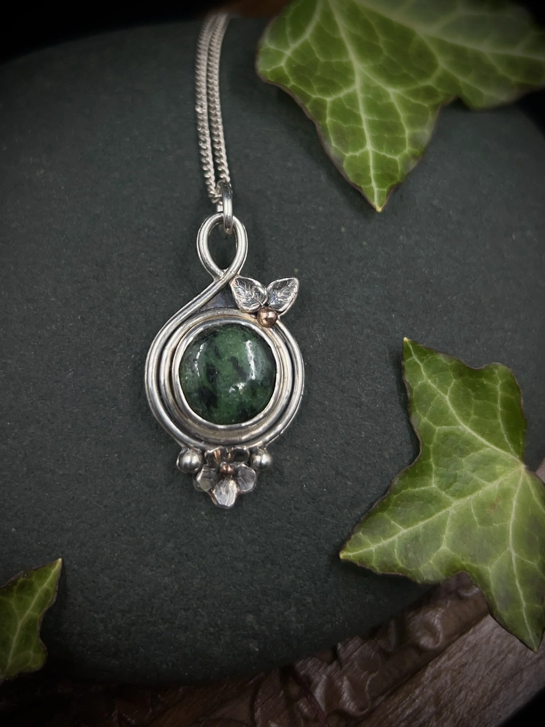 Silver and dark green round floral pendant with leaves, flowers and buds surrounding the stone displayed against an ivy background.