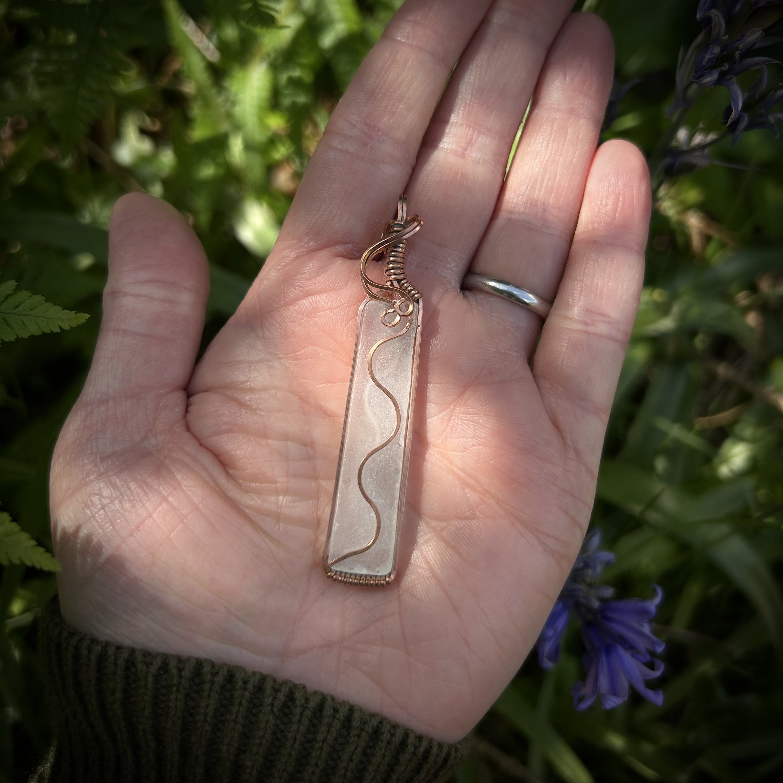 Model holding a long clear sea glass pendant on her palm. The piece is wrapped in copper with a copper wave on the front and back, highlighted by the dappled light.