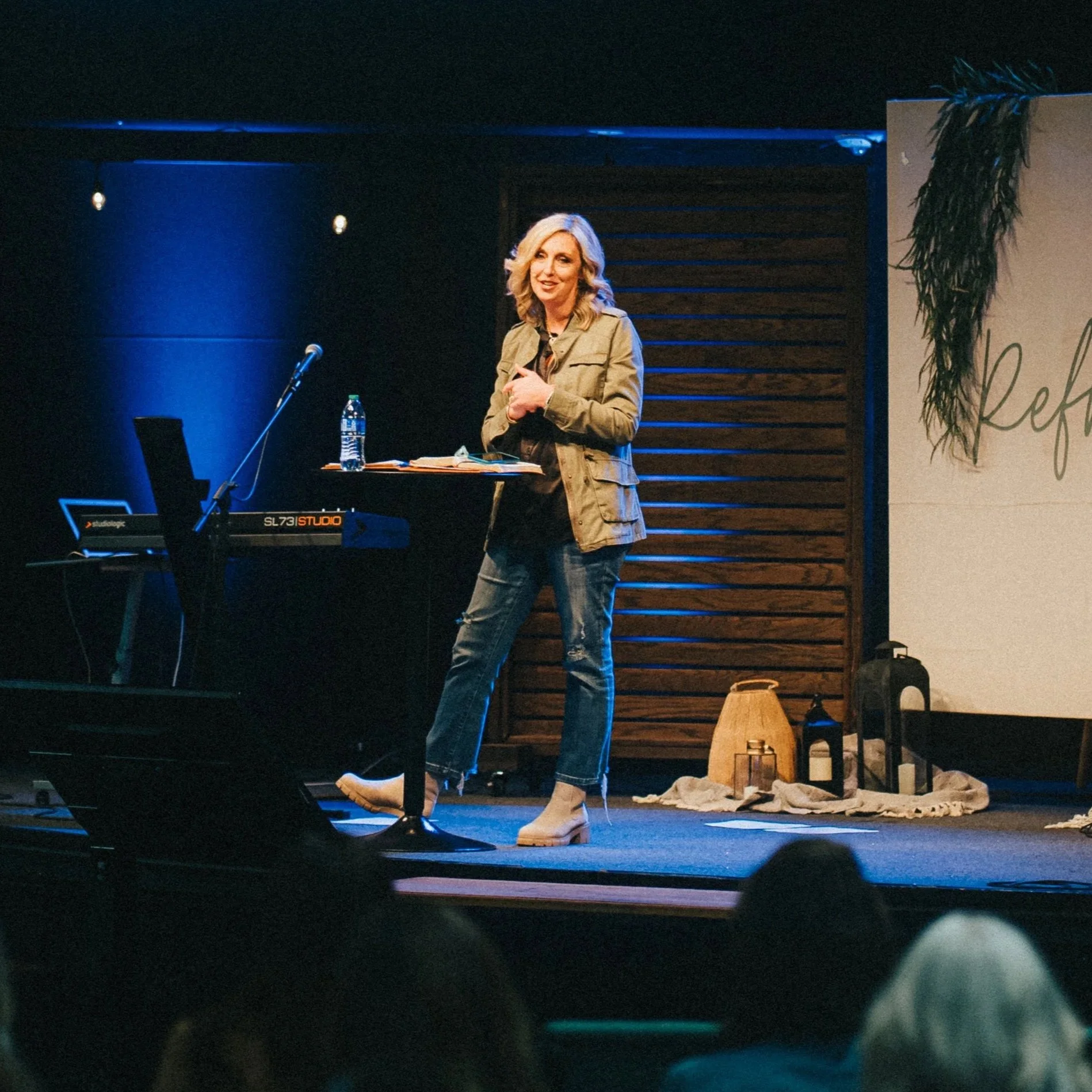 A woman with blonde hair standing on a stage, smiling, wearing a beige jacket, blue jeans, and cream-colored boots. Behind her, there is a wooden backdrop and a large sign with the word "Re" visible, along with a table holding papers, a water bottle, and a microphone. The stage is decorated with greenery and a cloth on the floor.