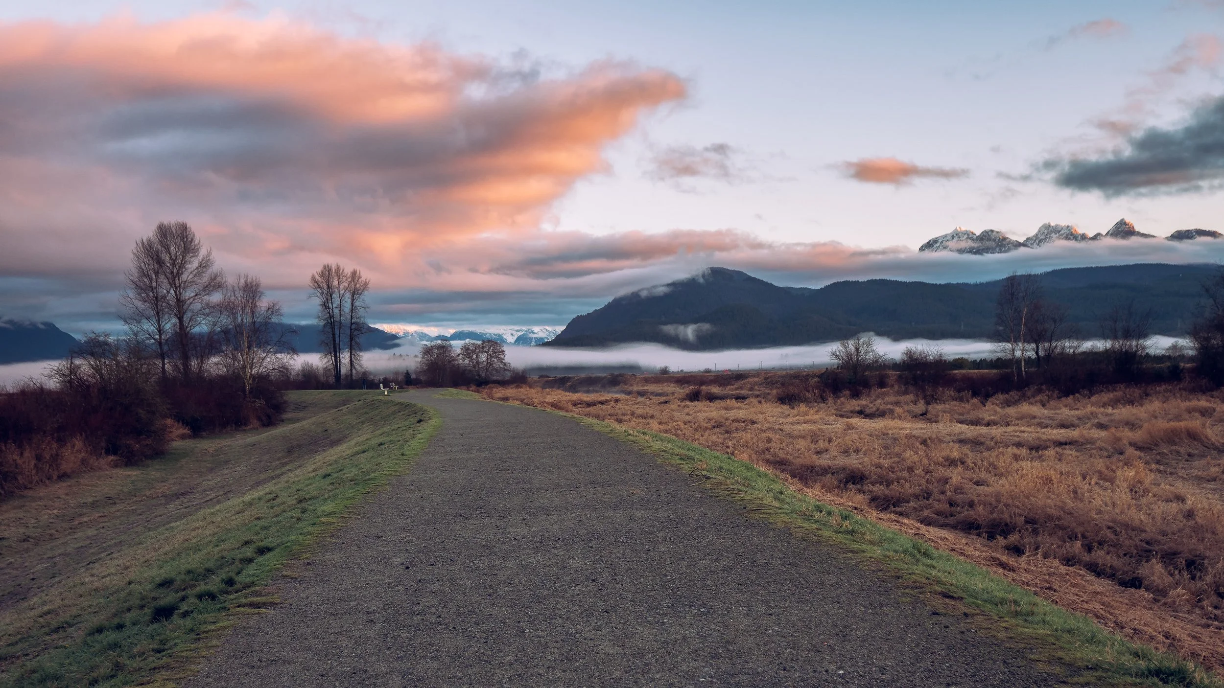 A gravel pathway winds through a rural landscape with mountains in the background, some trees on either side, and a colorful cloudy sky.
