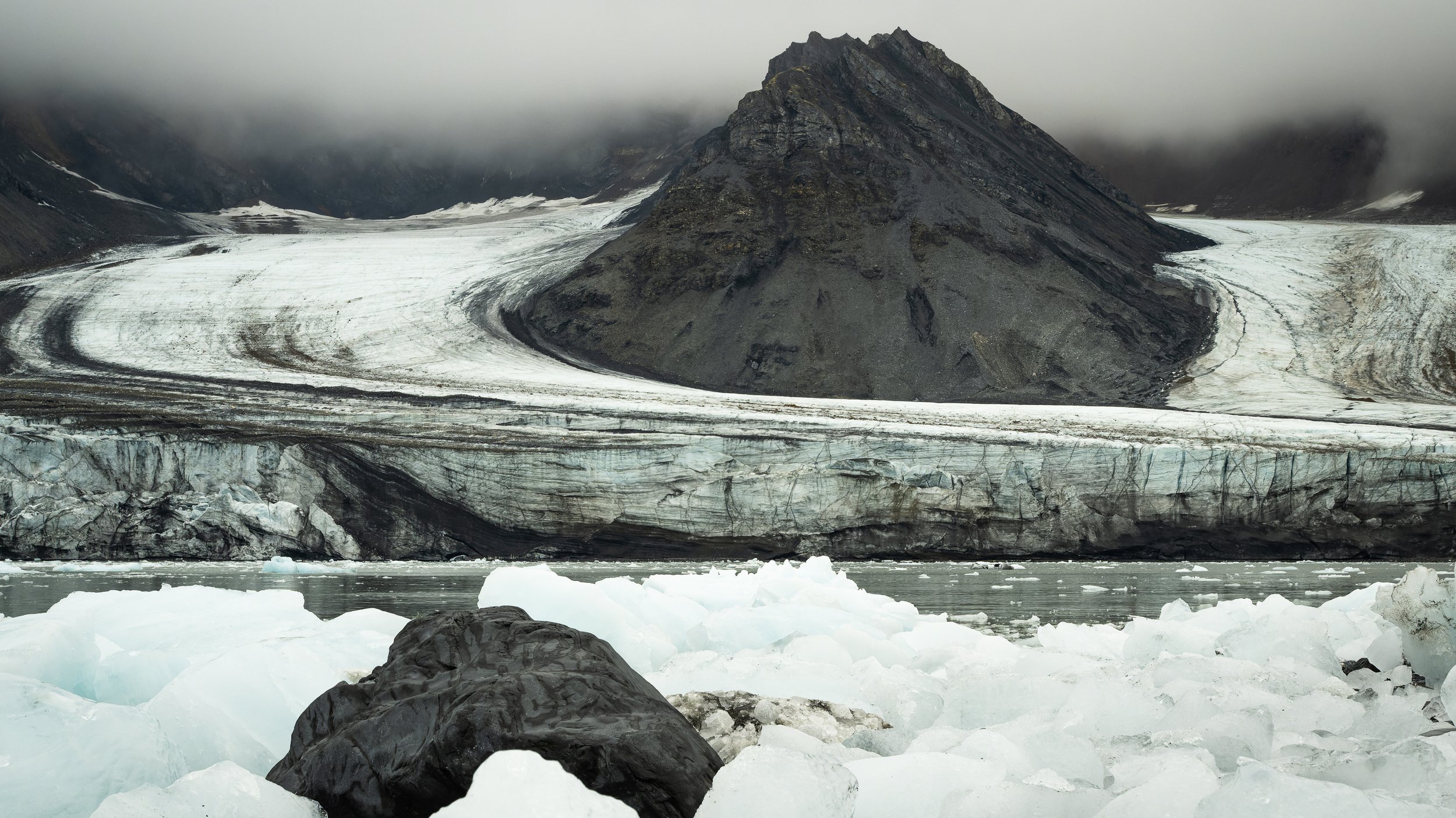 	Langue glaciaire serpentant entre des montagnes sombres, contraste entre la blancheur de la glace et la roche noire volcanique. Spitsberg
