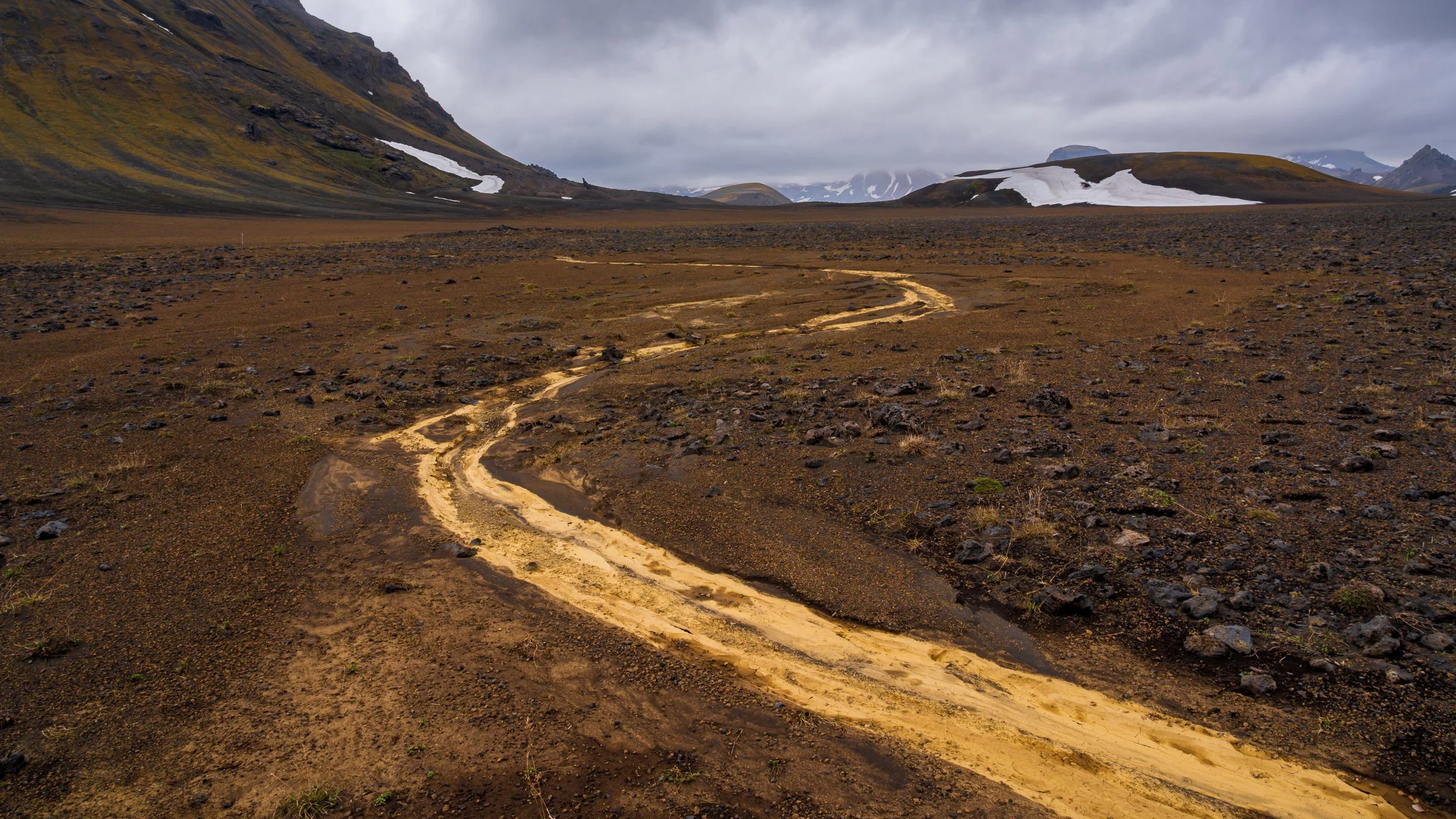 	Piste de terre serpentant à travers les hauts plateaux désertiques d'Islande, perspective vers les montagnes enneigées.