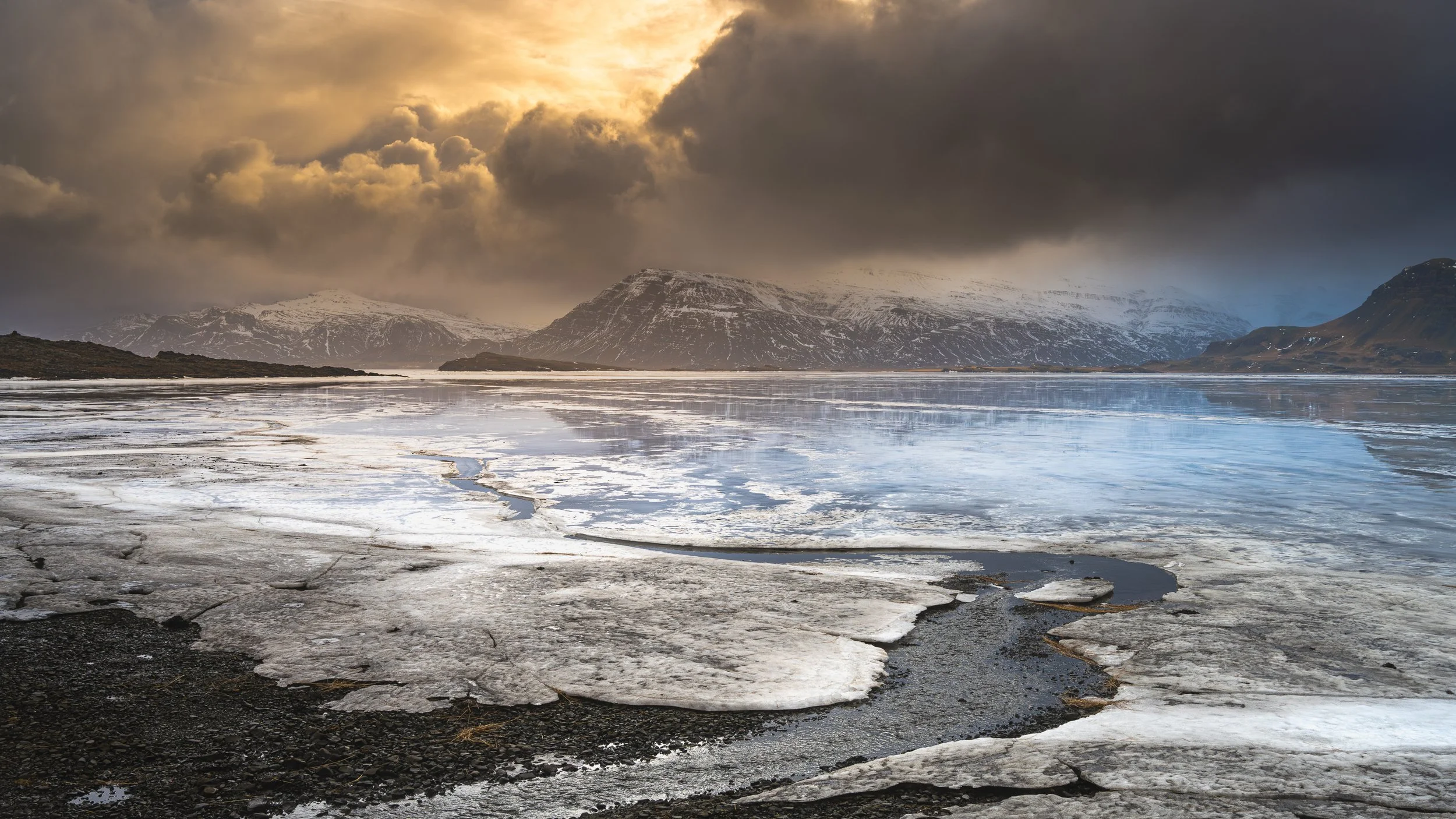 	Lumière dorée perçant les nuages au-dessus d'une plaine côtière islandaise, contraste entre terre rousse et ciel de tempête.