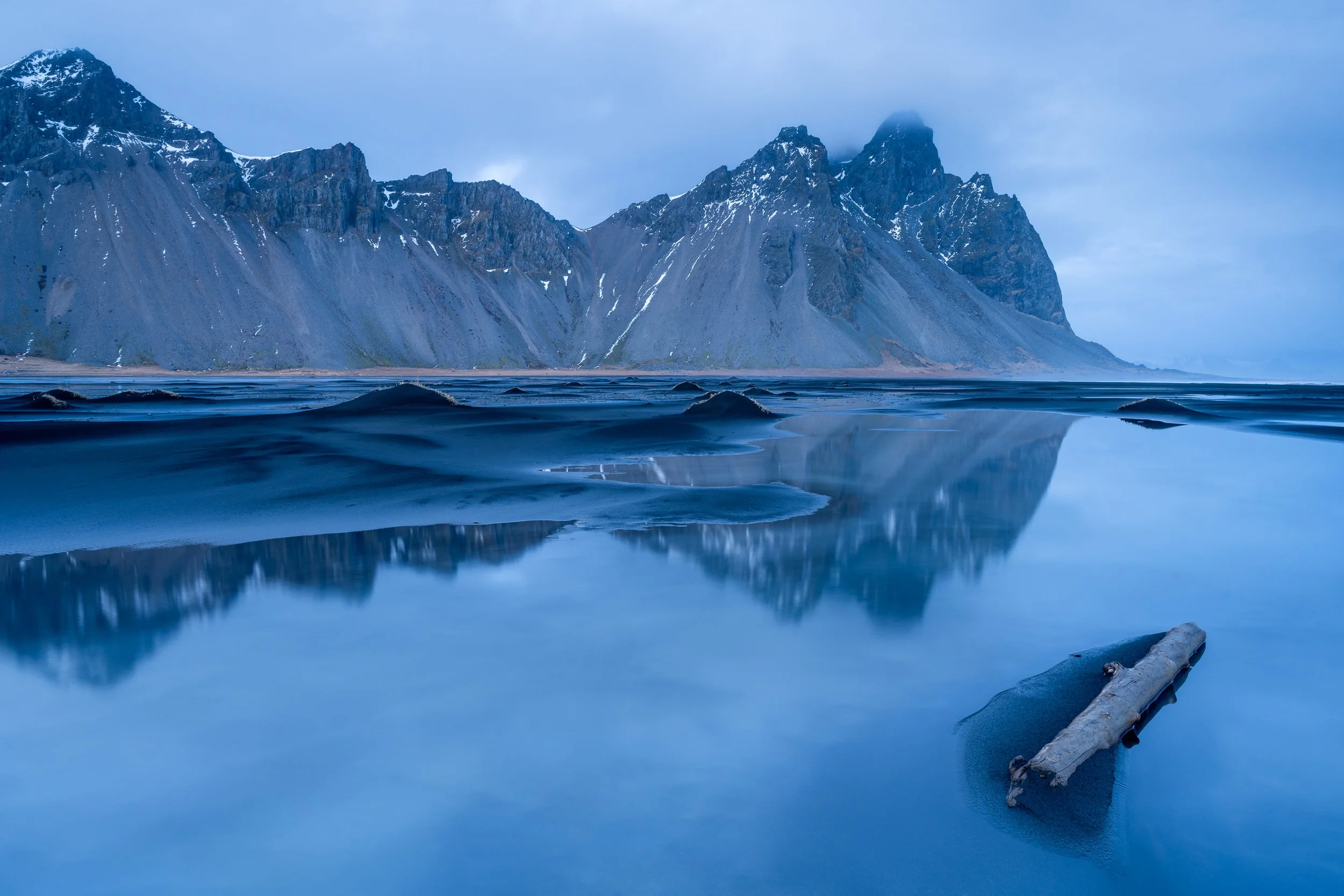 Reflet parfait de la montagne Vestrahorn dans l'eau d'une plage de sable noir en Islande, photographie de paysage en bleu profond.