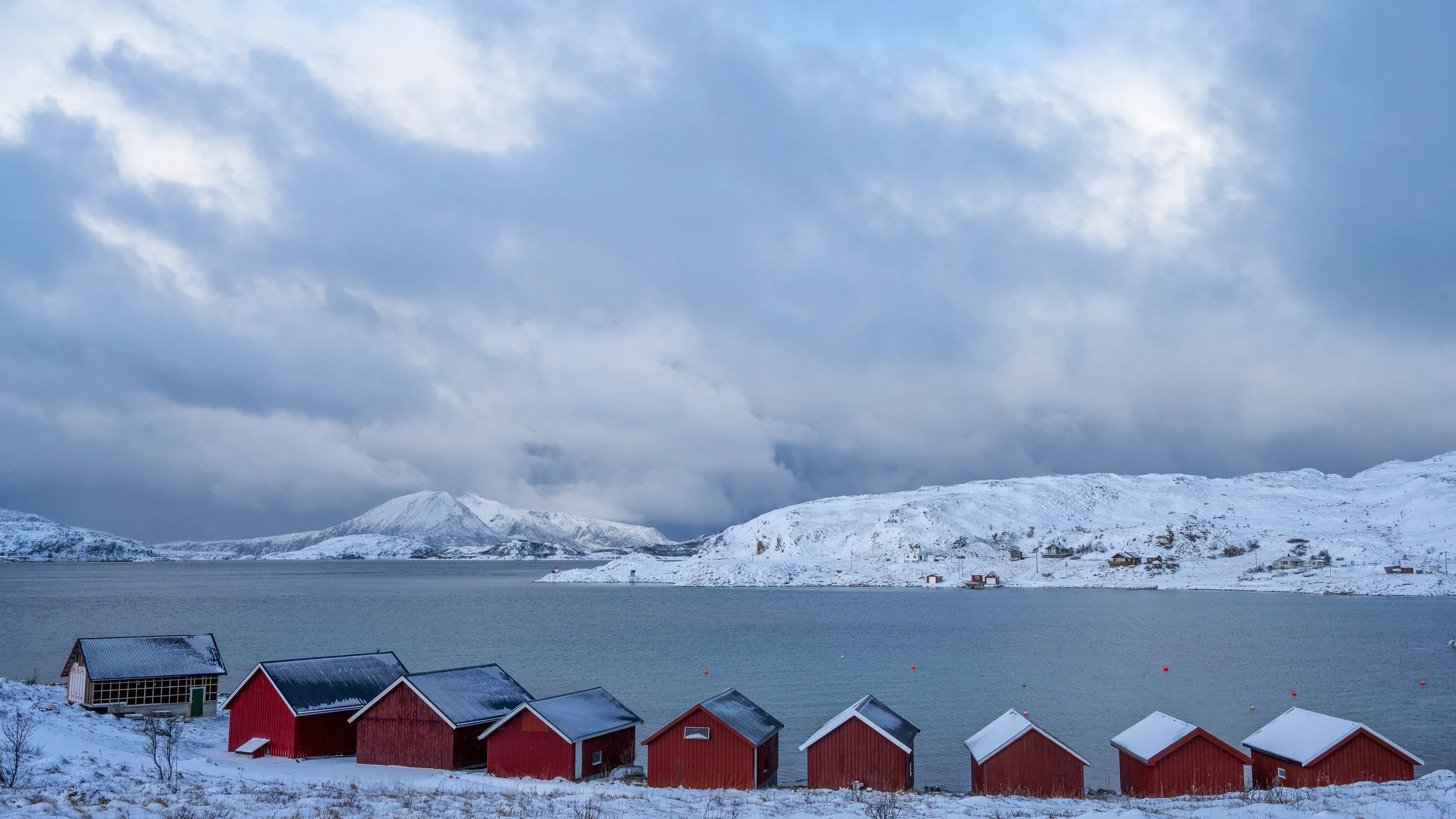 Alignement de cabanes de pêcheurs rouges en bord de fjord enneigé sous un ciel de tempête, ambiance Grand Nord. Norvège