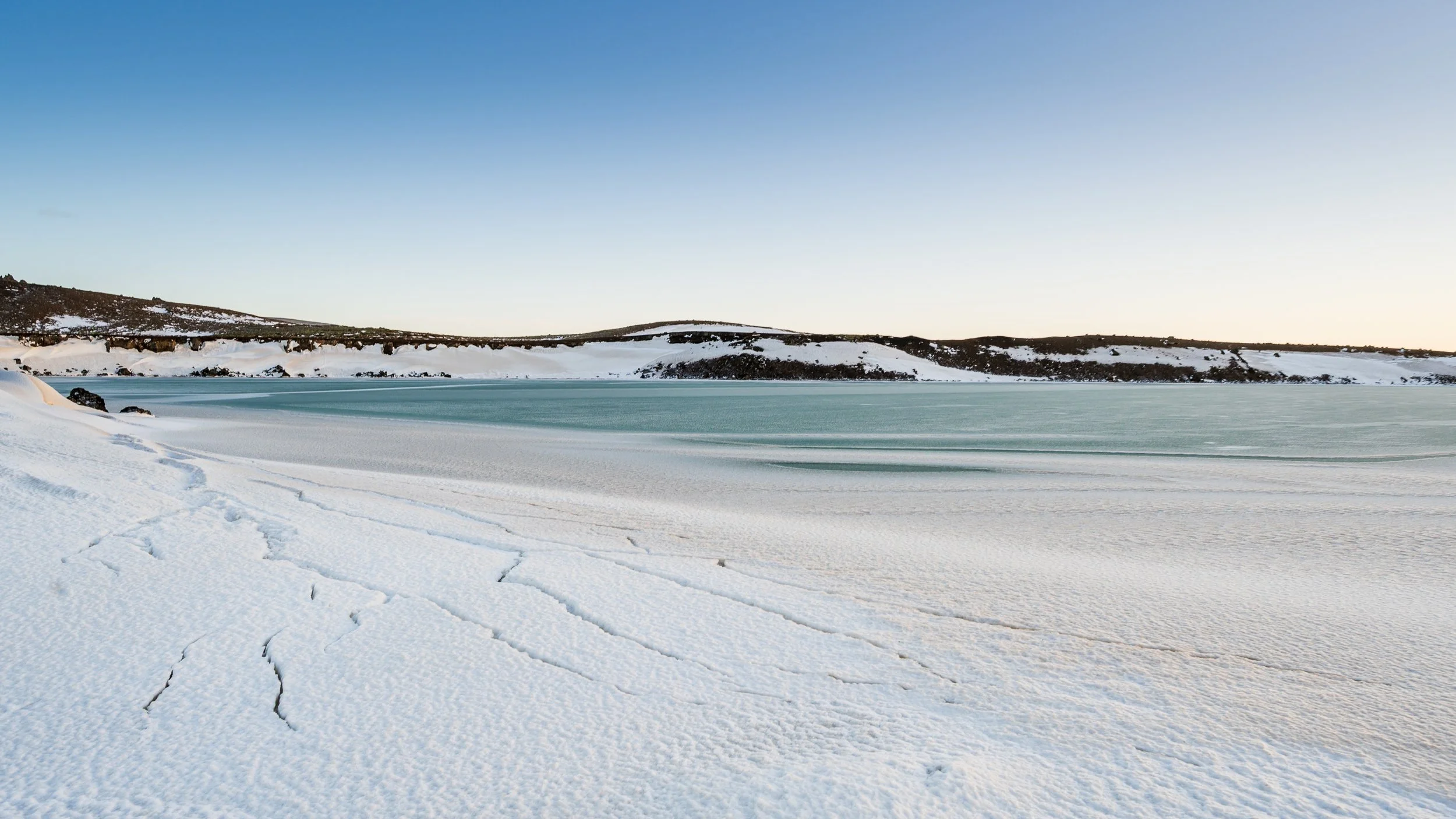 Rive enneigée d'un lac gelé sous un ciel bleu pâle, composition horizontale soulignant le calme et la solitude du Nord.