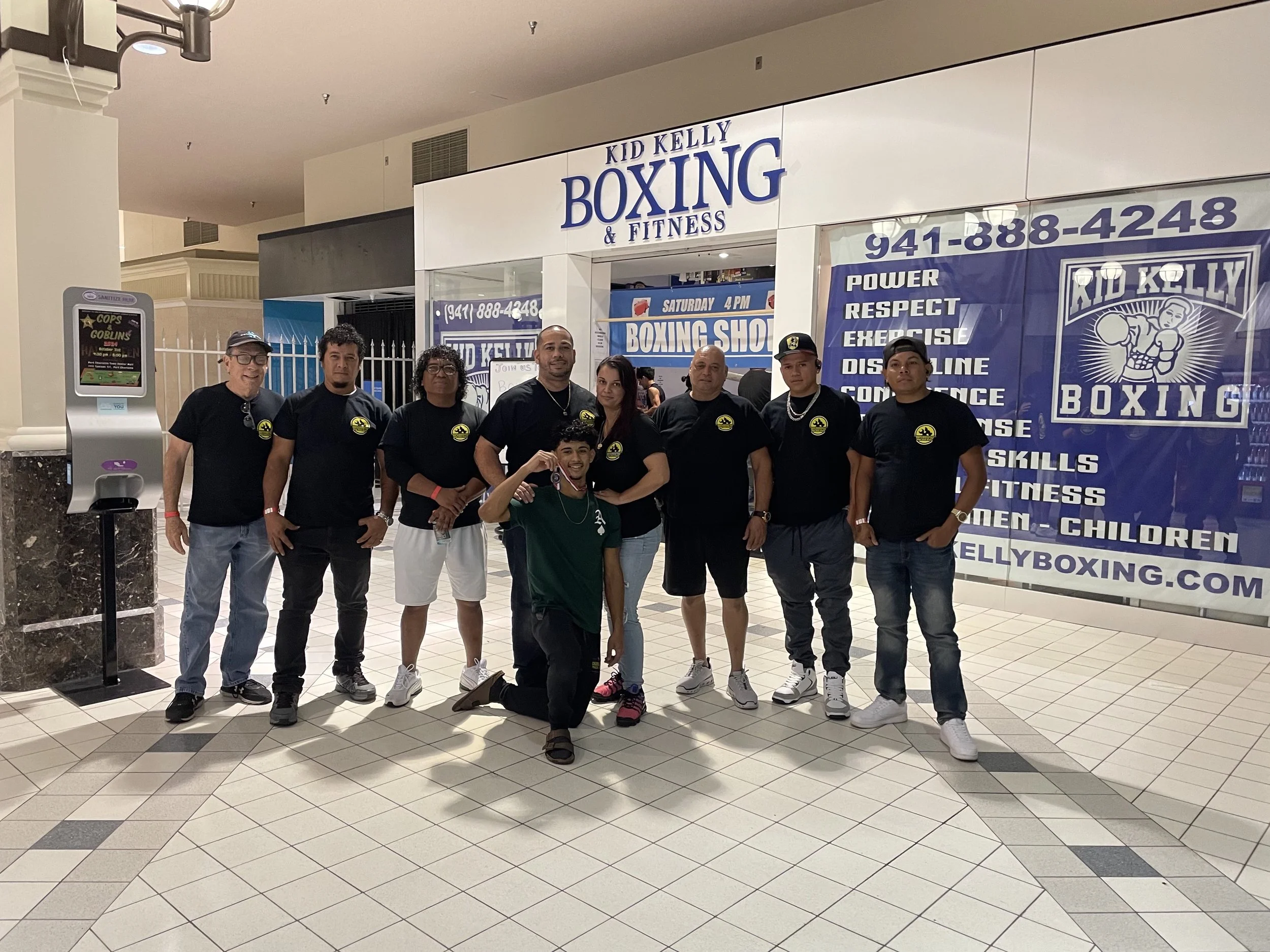 Group of seven people standing in front of a boxing and fitness gym entrance, with a large blue banner listing gym services and phone number, inside a shopping mall.