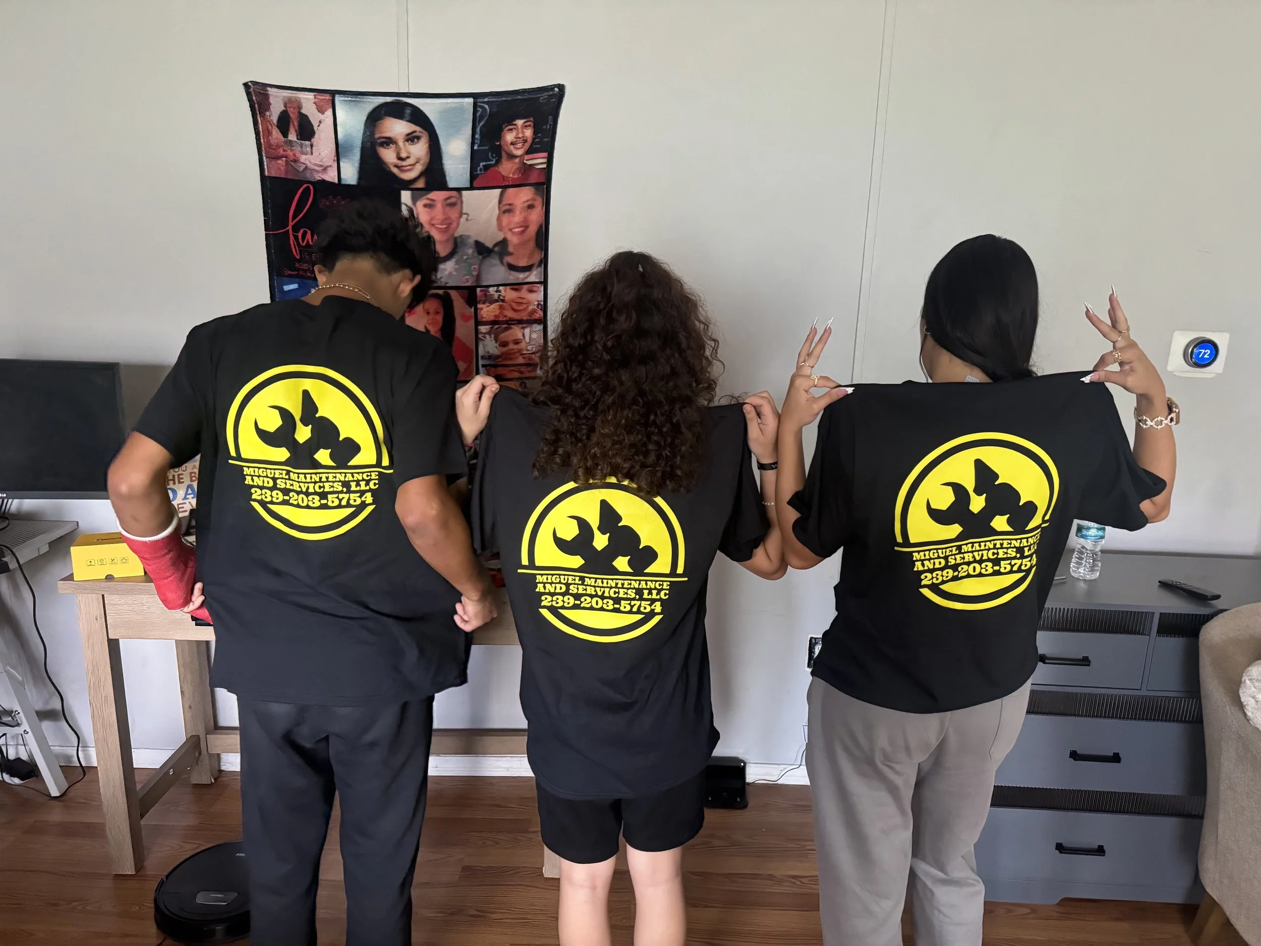 Three people are standing with their backs to the camera, facing a wall with a collage of photos of various individuals. They wear black shirts with yellow logos and contact information for a maintenance and cleaning service. One person has curly hair, another has dark straight hair, and the third has short dark hair.