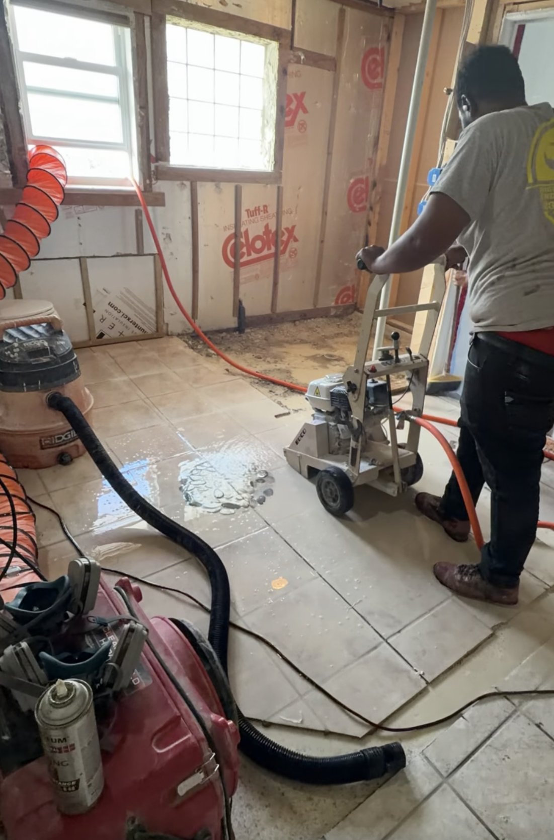 A person using a tile saw to cut tiles in a room under renovation with exposed wall framing and insulation. Construction tools and equipment are visible, including a vacuum, air compressor, and various cords.