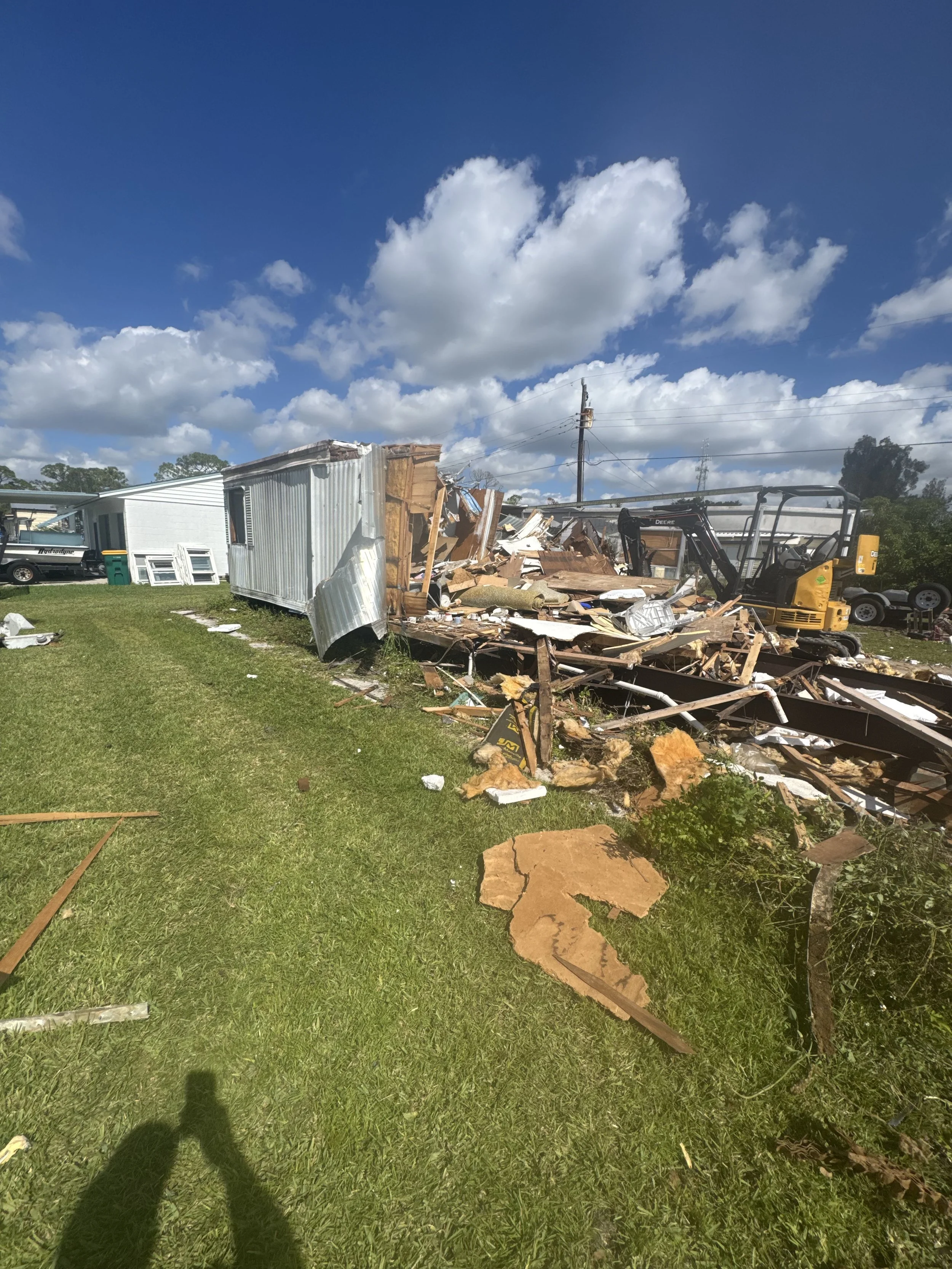 A destroyed building with debris scattered on the ground, including wood and metal, next to a small yellow excavator, under a blue sky with clouds.