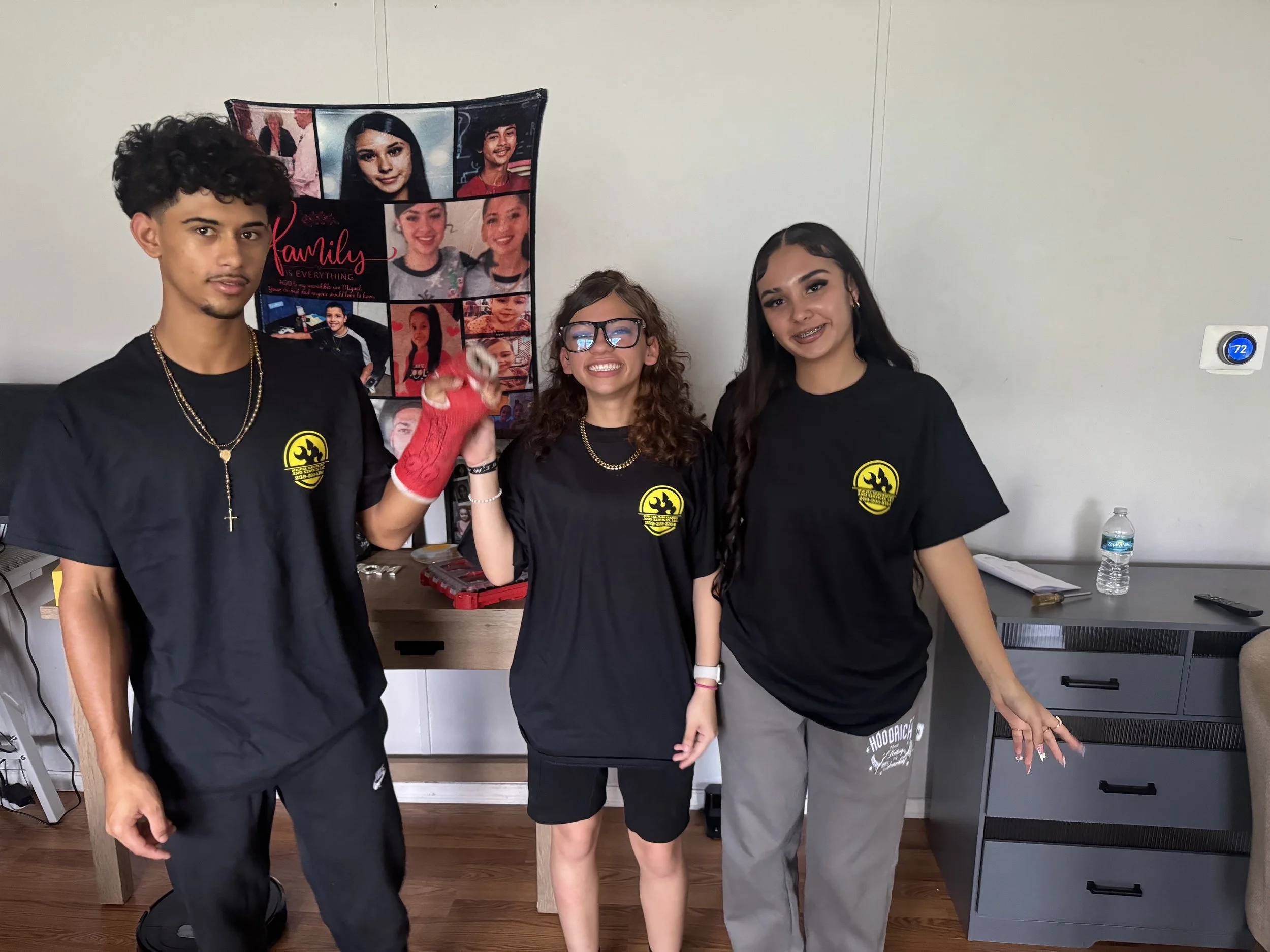 Three teenagers standing indoors in front of a photo collage. All three are wearing black t-shirts with a yellow logo. The boy on the left has curly dark hair, a necklace, and light skin. The girl in the middle has curly brown hair, glasses, and is smiling. The girl on the right has long dark hair, braces, and is looking slightly to the side. There is a water bottle on a desk and a thermostat on the wall.