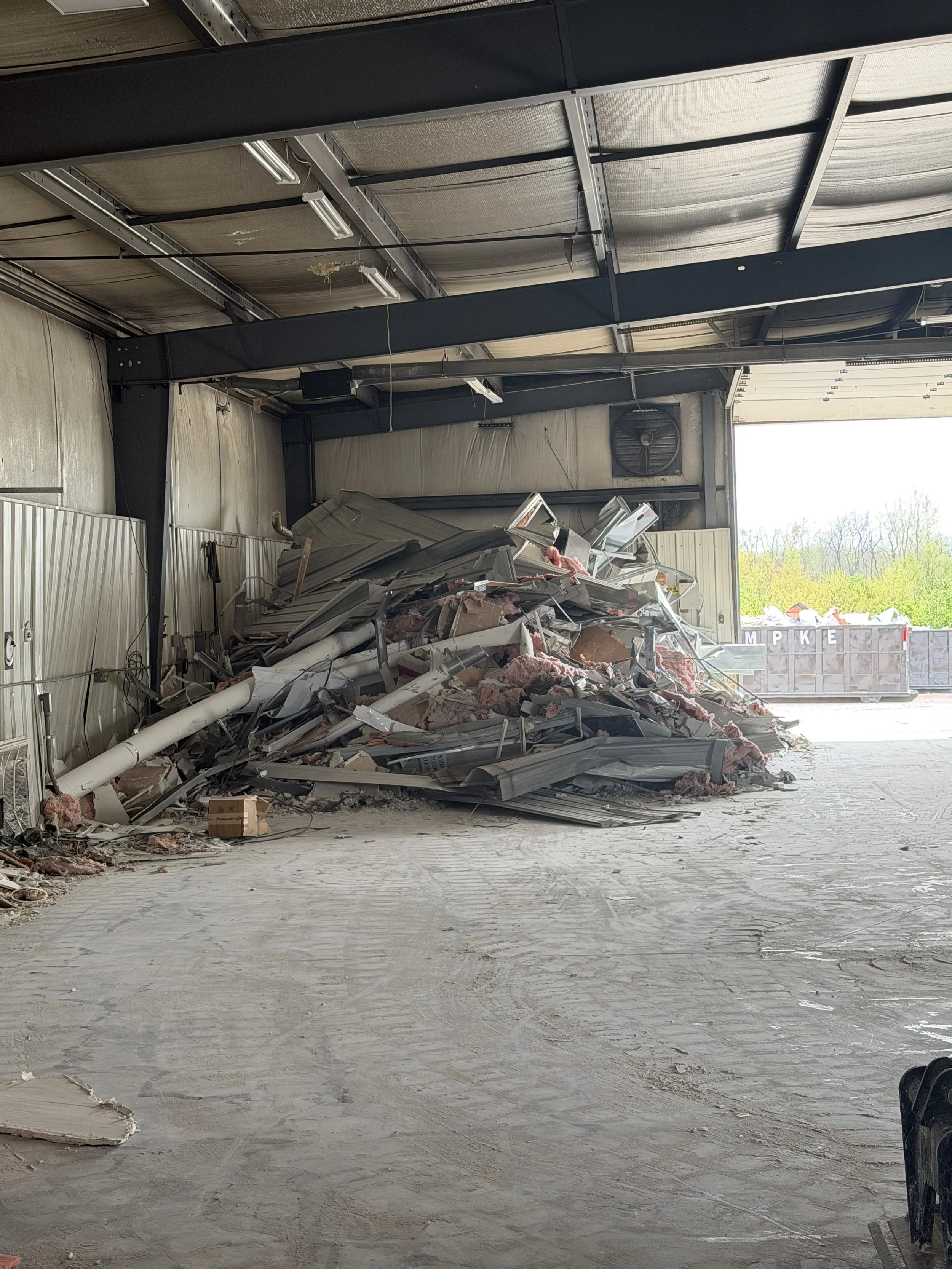 The image shows the interior of a large industrial or warehouse building with a high ceiling. There is debris, construction materials, and insulation scattered on the floor near the open garage door, with some metal and piping mixed in. Outside, there are trees and a truck container visible.
