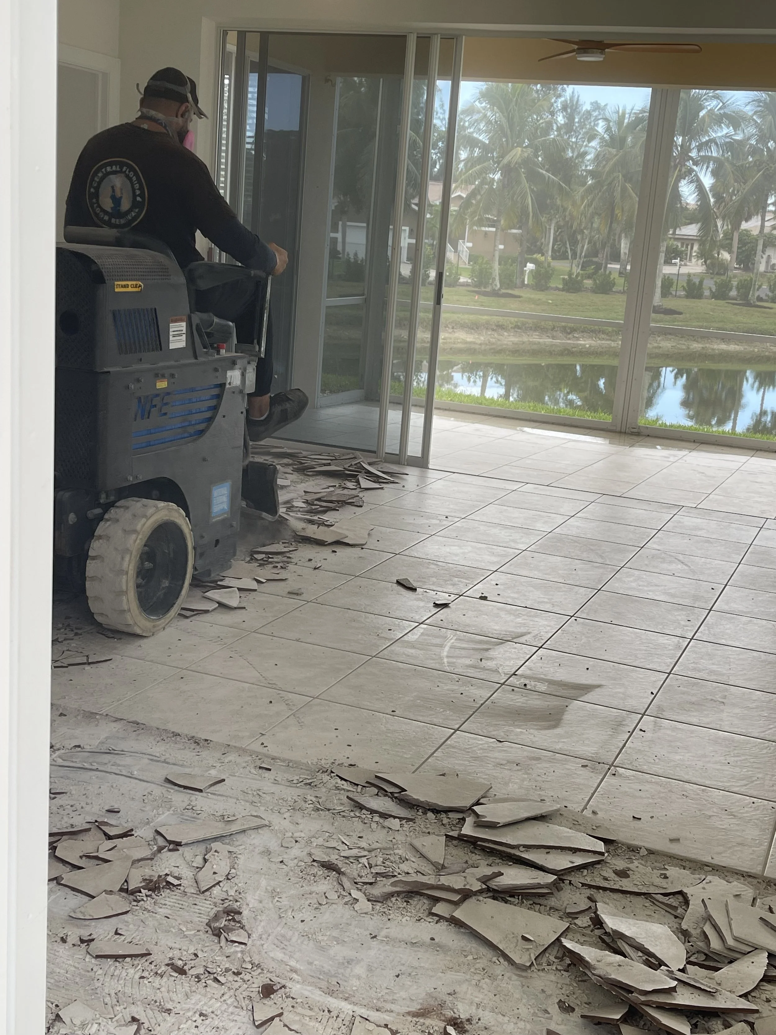 A person operating a floor scraper machine in a room with broken tiles, with an outdoor view of palm trees through large glass doors.