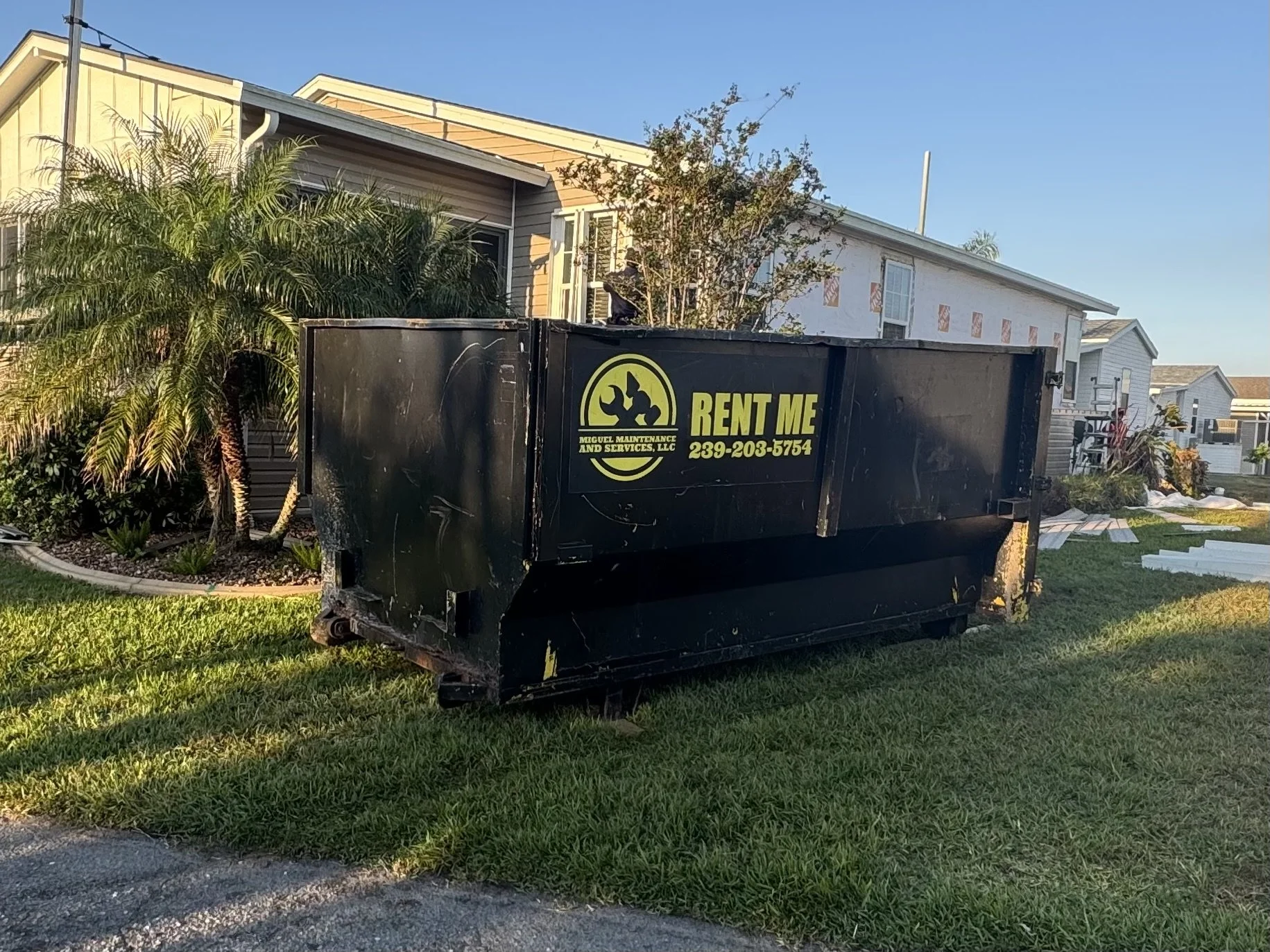 A black dumpster with a yellow and black sign that reads 'Miguel Maintenance and Services, LLC' and a phone number, situated on a grassy yard in front of a house under construction or renovation.