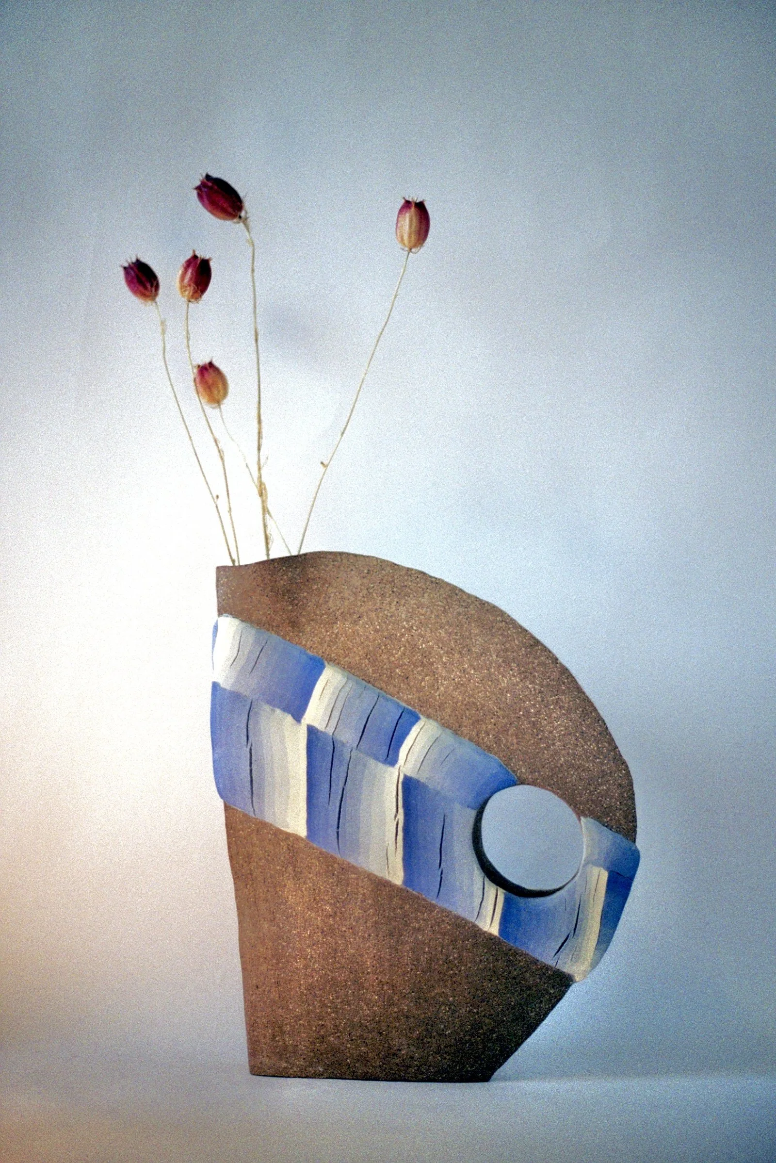 A modern art sculpture features a brown textured oval base with a blue and white striped band wrapping around it, a circle at the front, and tall dried pink flower stems emerging from the top.