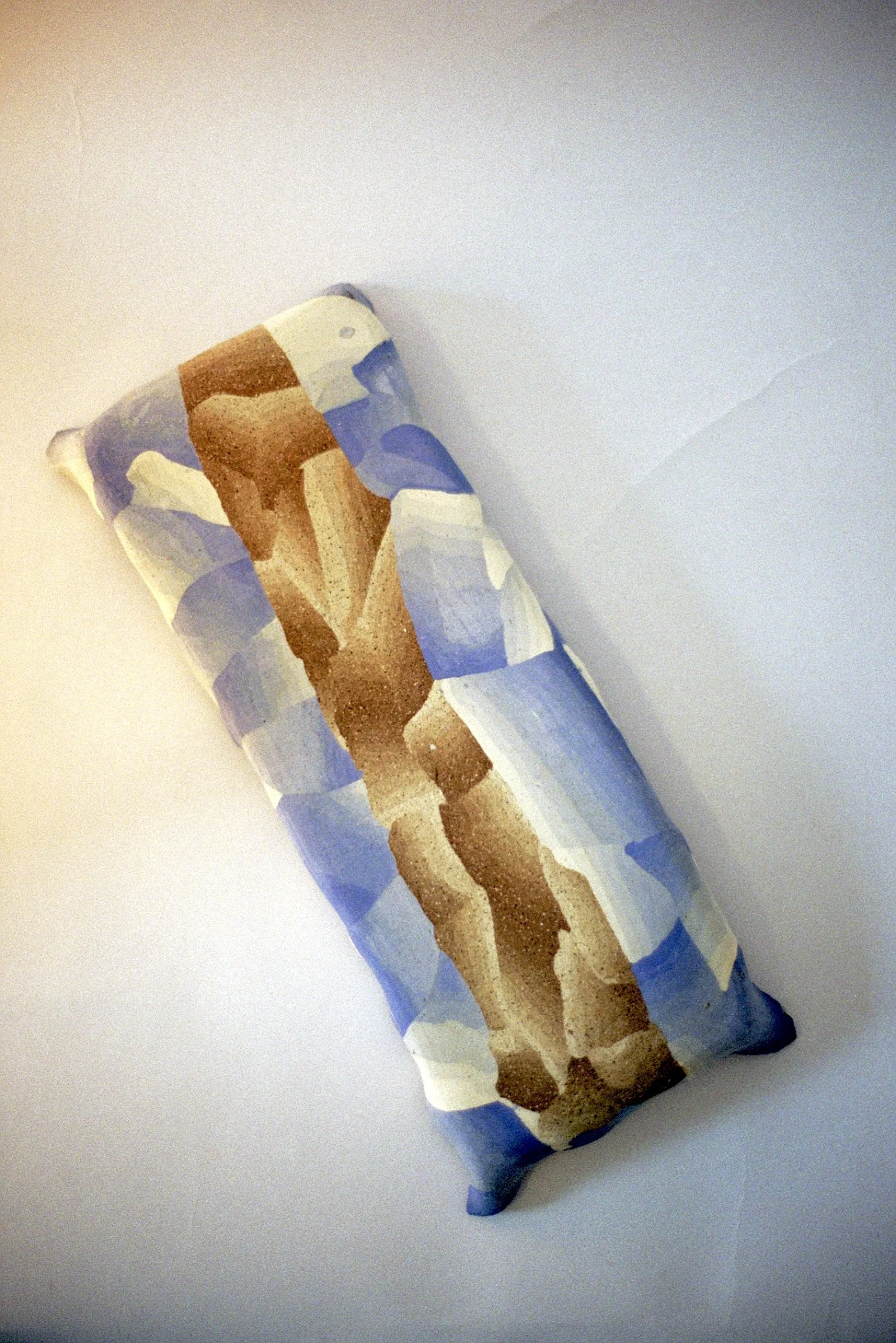 A rectangular pillow with a geometric and abstract design in shades of brown, tan, cream, and blue, resting on a white surface.