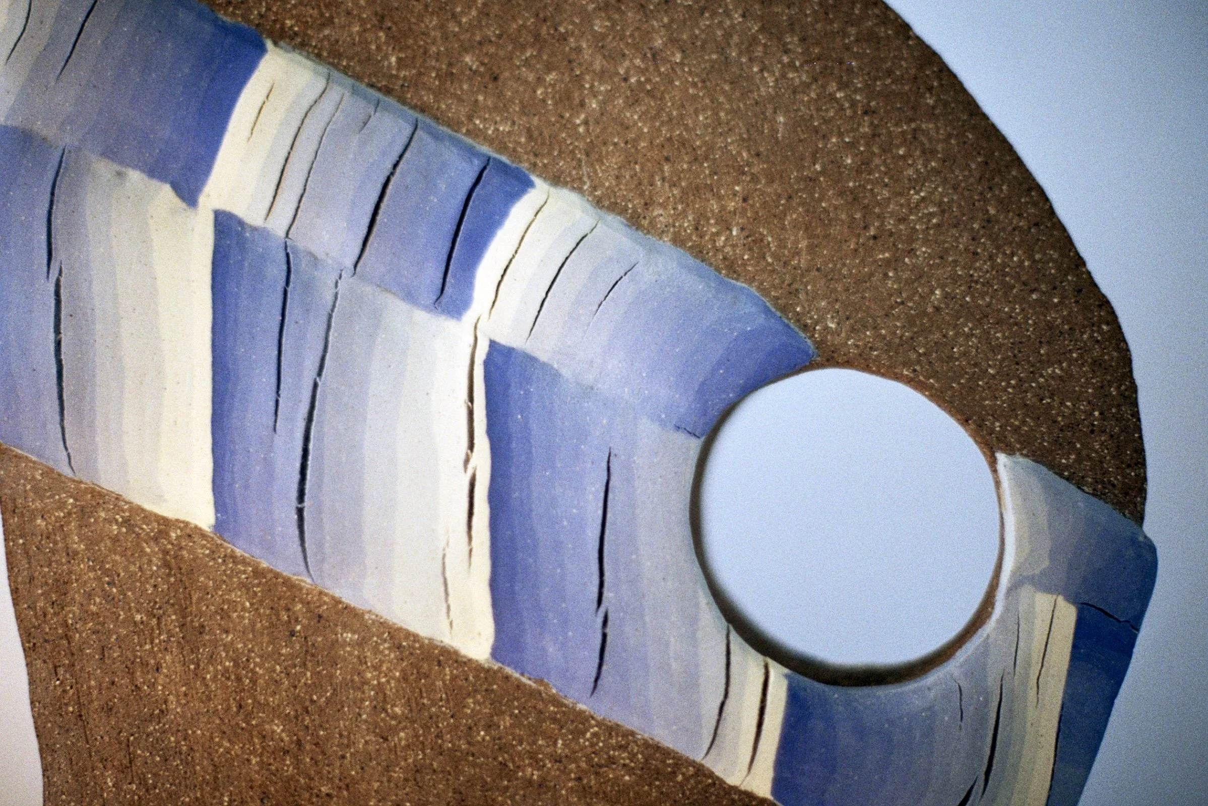 Fragment of a ceramic sculpture with blue, white, and beige stripes and a circular hole, resting on a textured brown surface against a clear blue sky.