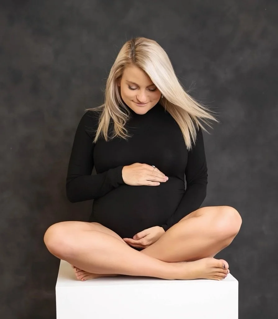 A pregnant woman with blonde hair, wearing a black long-sleeved top, is sitting cross-legged on a white platform against a dark background. She is gently holding her belly with one hand and looking down at it.