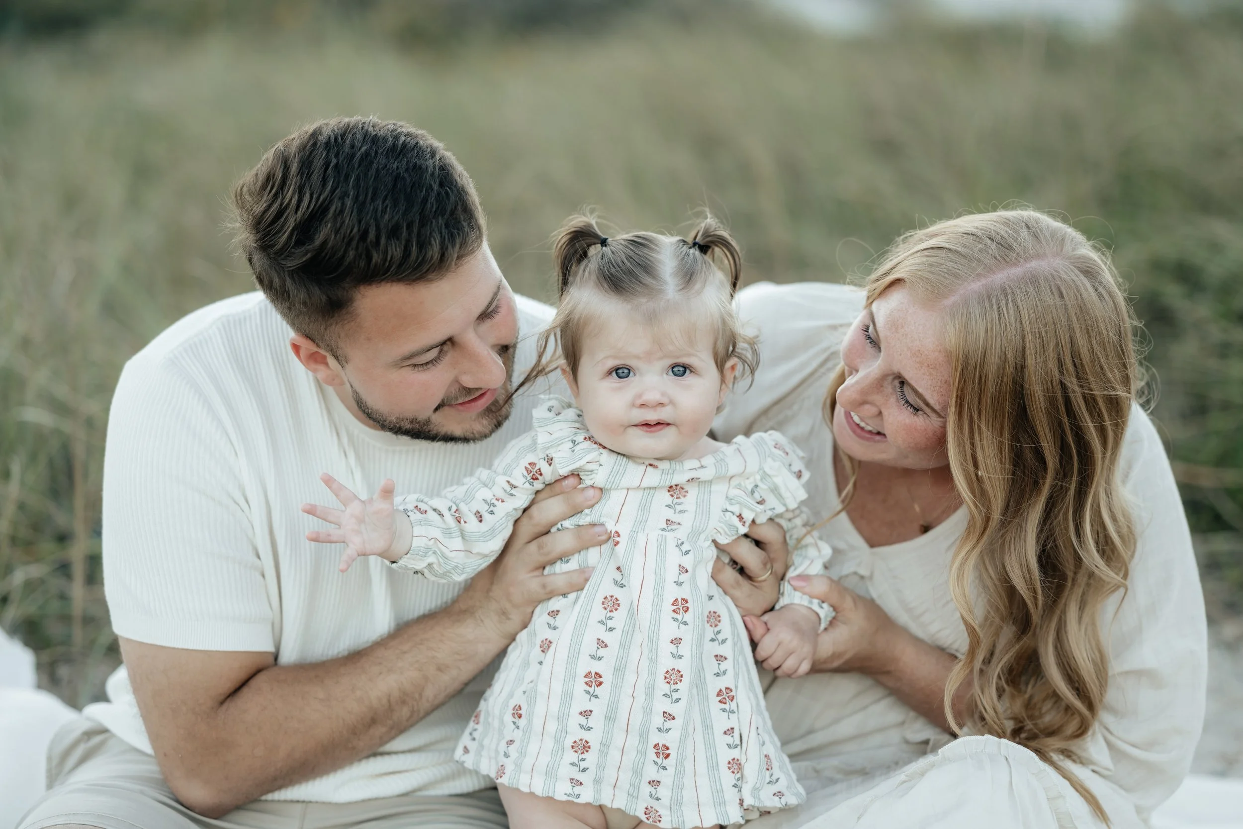 mom and dad with their daughter sitting in the grass at the beach in jacksonville florida