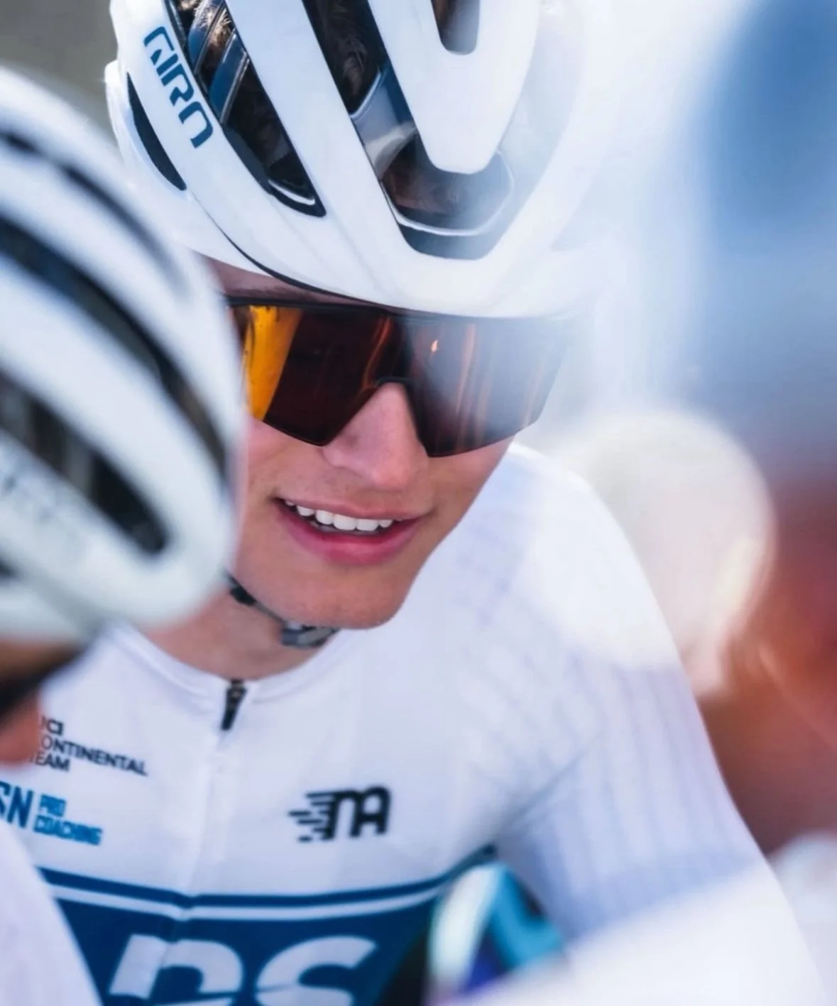A male cyclist wearing a white helmet, sunglasses, a black and green cycling jersey, white socks, and blue cycling shoes is bending down to adjust his shoes on an outdoor road.