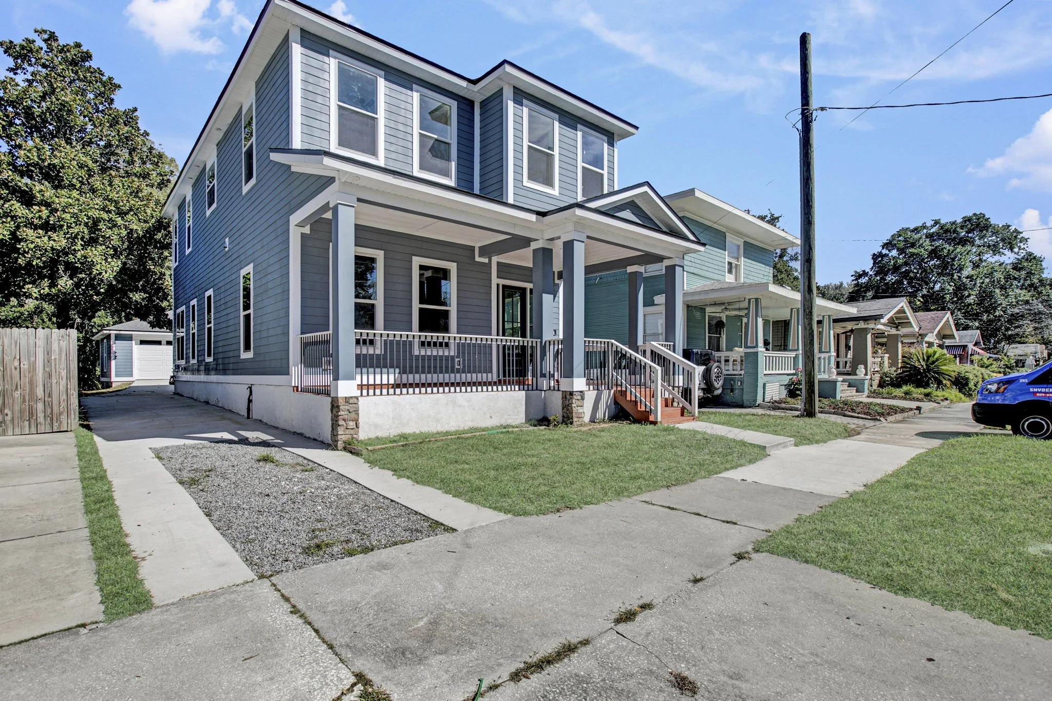A row of modern, two-story houses with front porches, painted in shades of blue, under a clear blue sky. The houses are situated along a concrete sidewalk with a grassy yard area.
