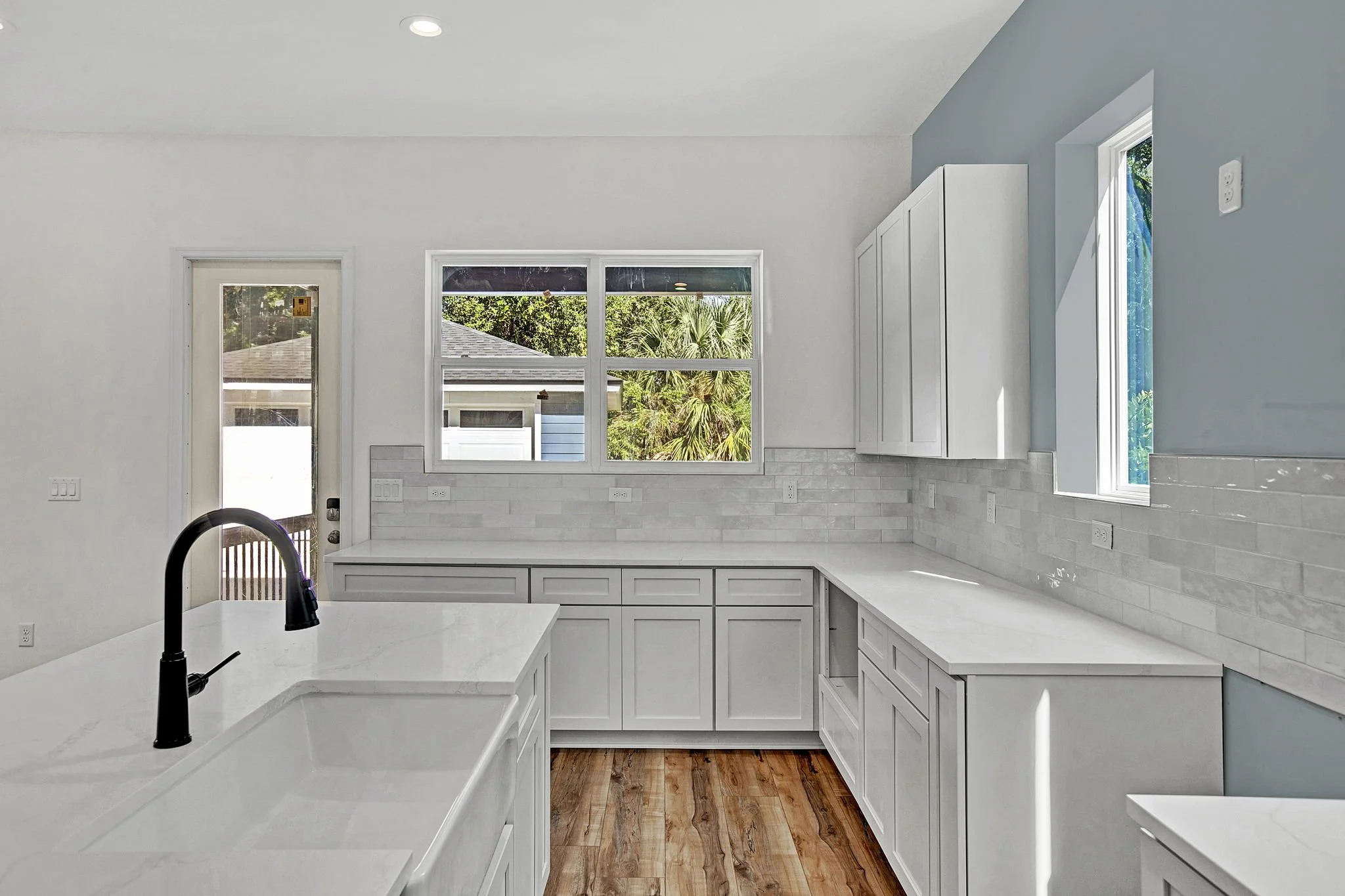 Empty kitchen with white cabinets, white subway tile backsplash, large windows, and a black faucet over a white sink.