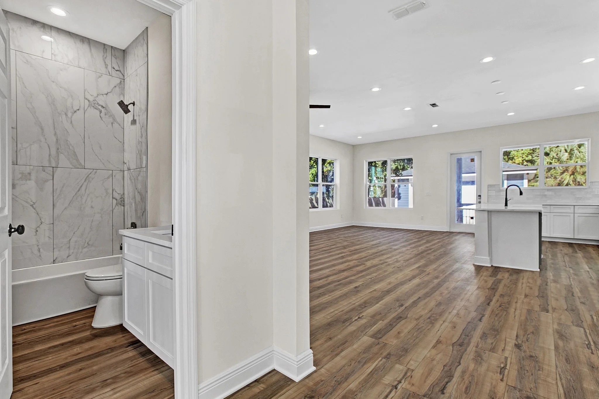 Empty living room and kitchen area in a modern house with wood flooring, white walls, large windows, and a view of a backyard. Part of the bathroom with a marble-tiled shower and toilet is visible on the left.