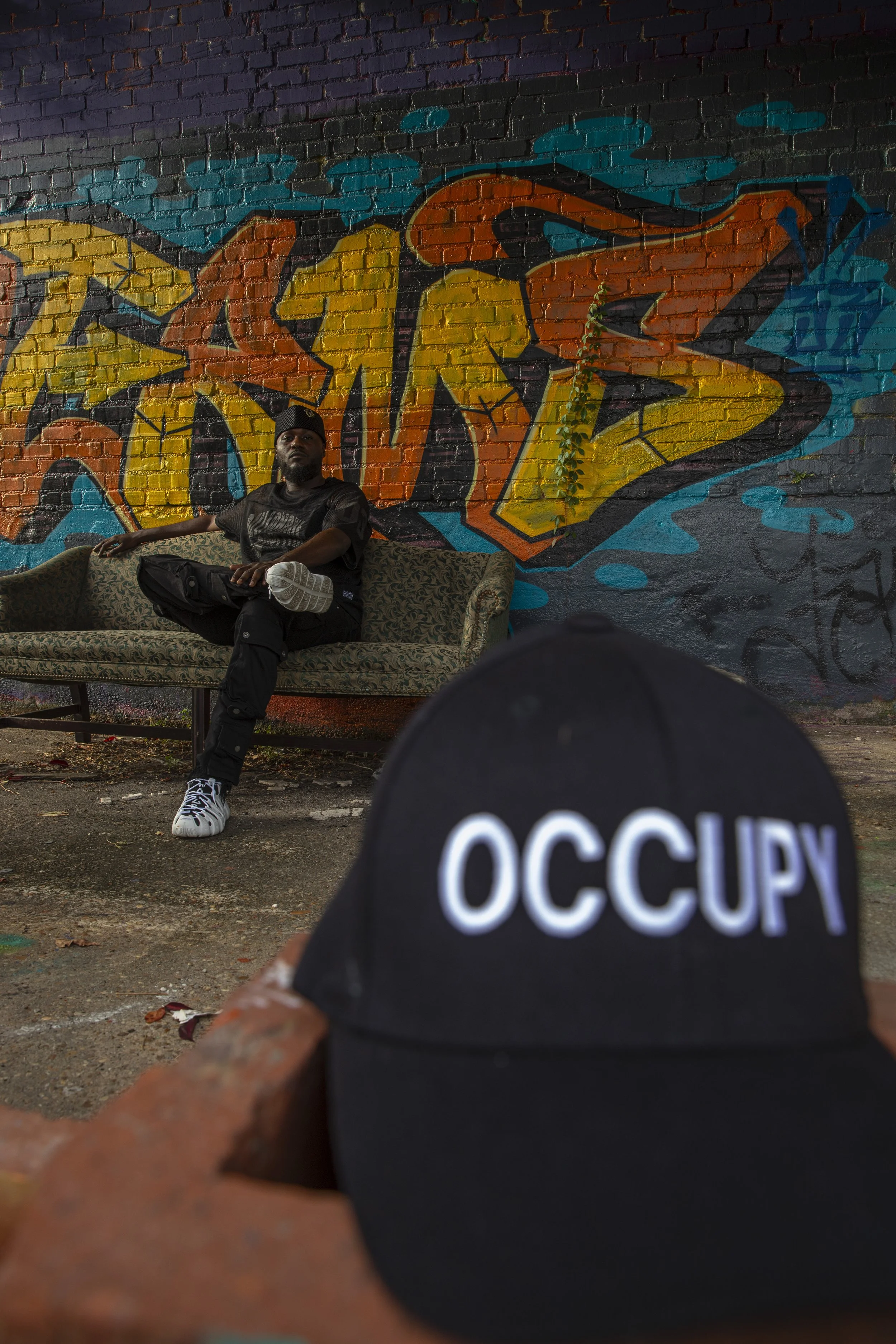 A man sitting on a vintage couch with legs crossed in front of a colorful graffiti mural on a brick wall, with a black cap and cap with 'OCCUPY' written on it in the foreground.