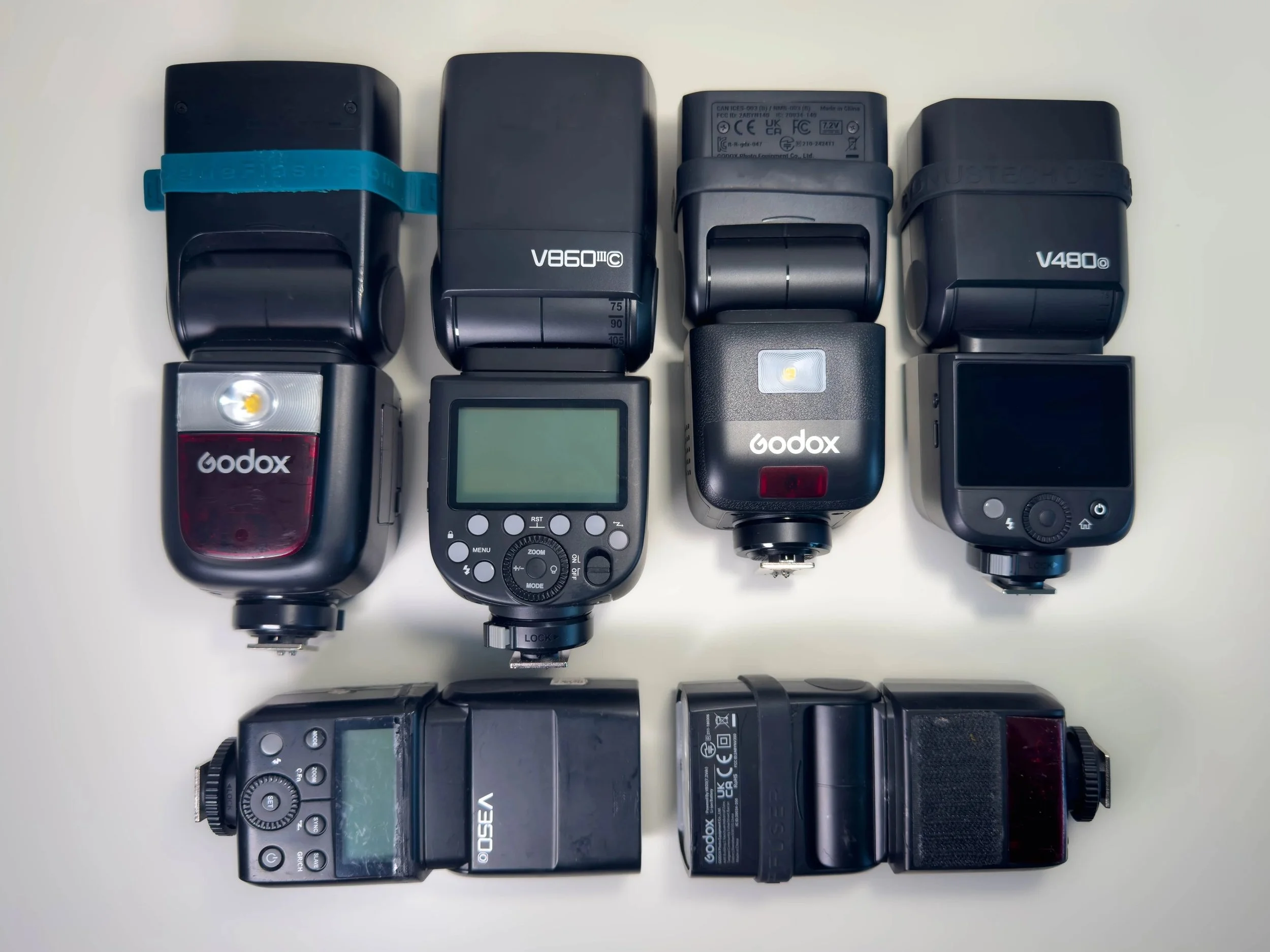 Collection of camera flashes and speedlights of various brands and models on a white surface.