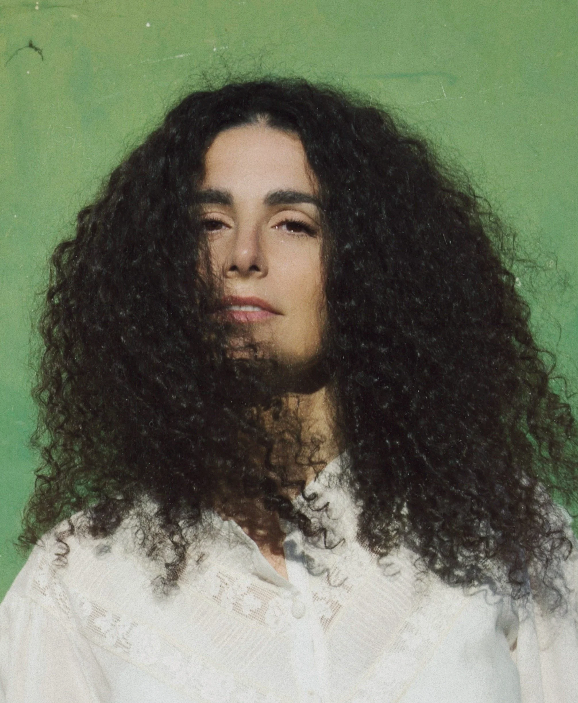 A woman with voluminous curly black hair, light skin, wearing a white top, standing in front of a green background.