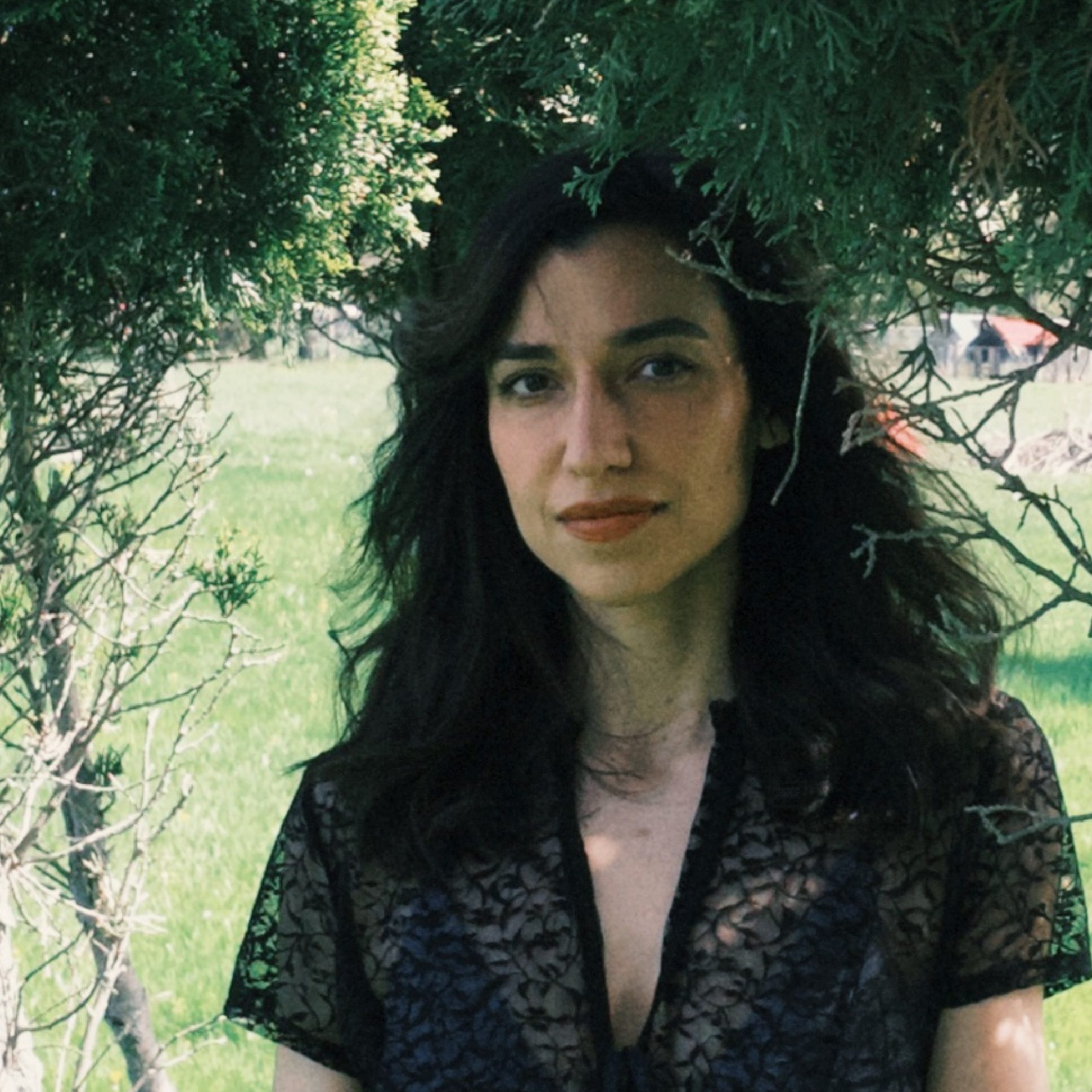A woman with dark wavy hair standing outdoors under a green tree, wearing a black lace top, with a blurred background of a grass field and structures.