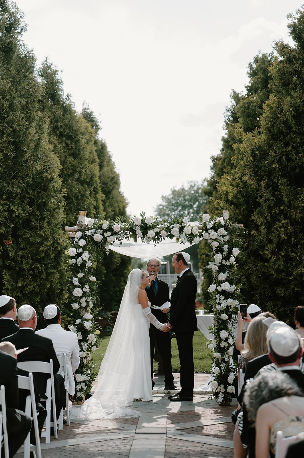 A wedding ceremony outdoors with a bride and groom holding hands in front of an officiant, under a floral archway. Guests seated on white chairs are watching, many wearing kippahs. The setting includes tall greenery and a partly cloudy sky.