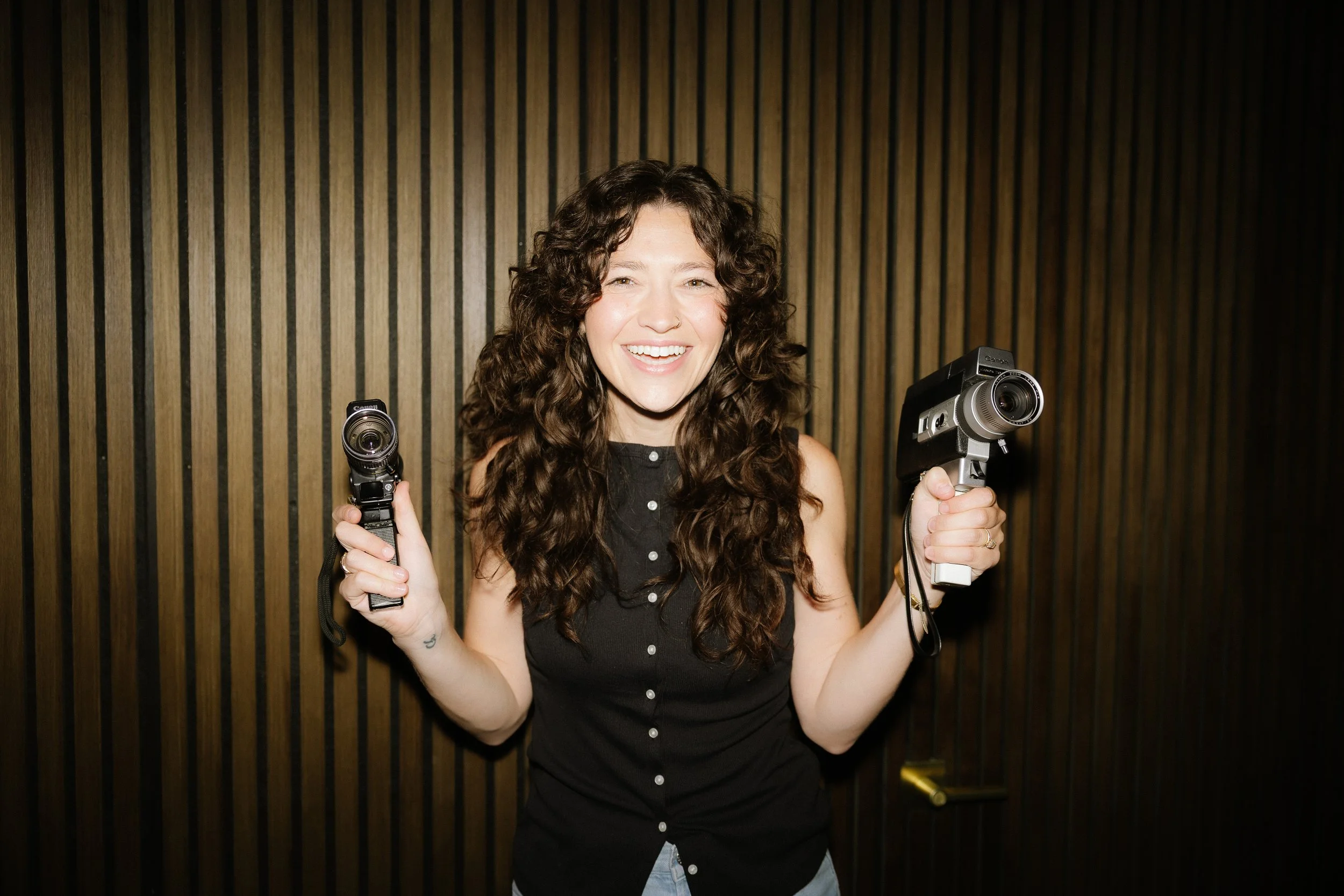 A young woman with long curly brown hair, smiling and holding two vintage camcorders, standing against a wooden vertical slat wall.