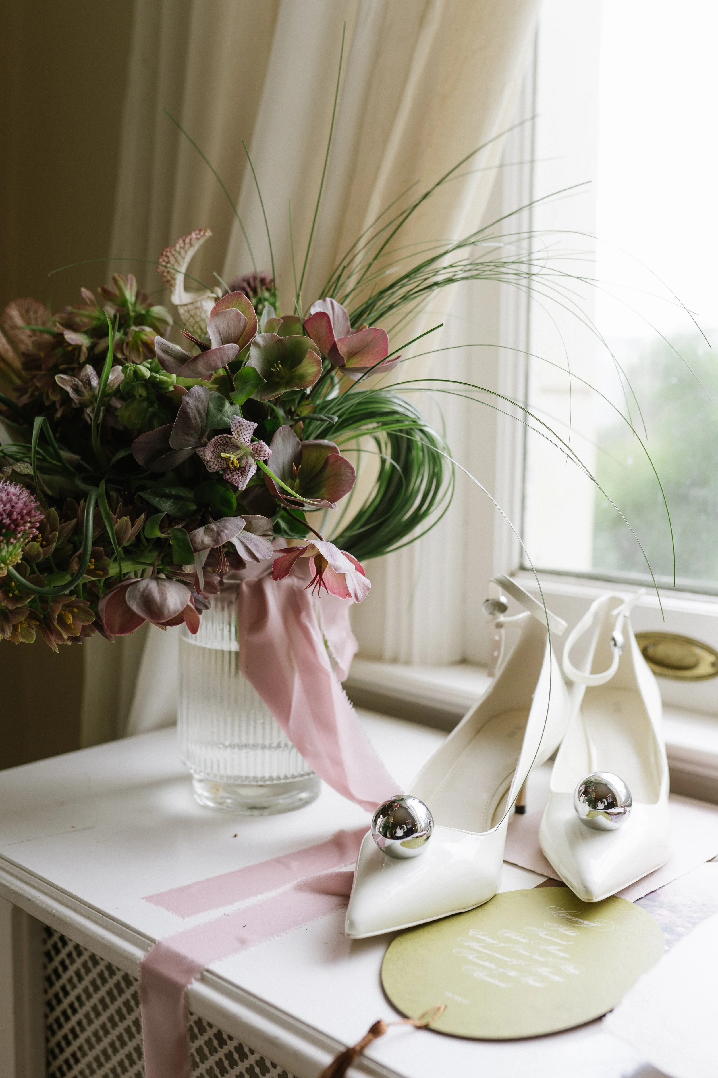 Image of bridal details on a window sill included an organic bouquet of purple and greens next to white shoes with silver embellishments