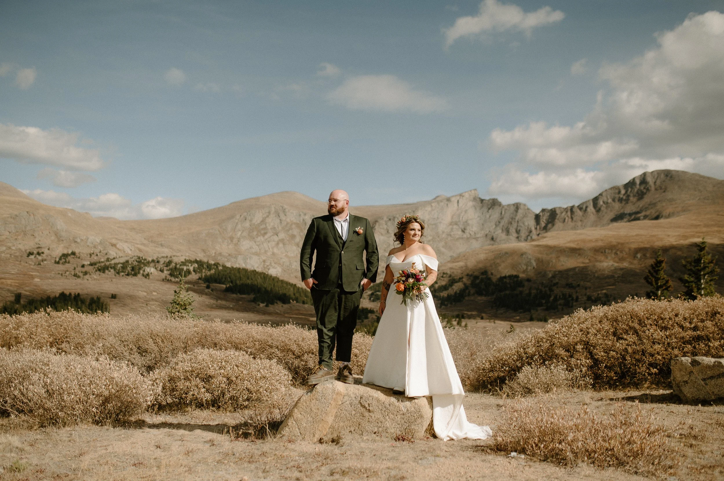 A bride and groom walking outdoors in a mountainous landscape on their wedding day, with a clear sky and scattered clouds overhead.
