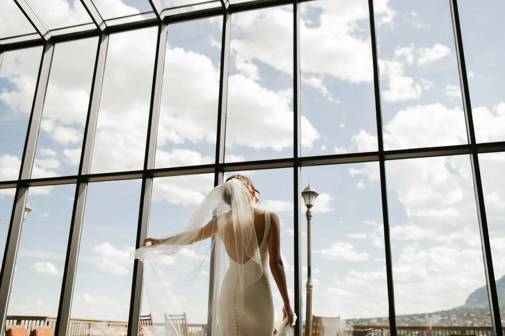 A bride in a white wedding dress with a veil, standing in front of large glass windows with an outdoor view of a sky with clouds and mountains.
