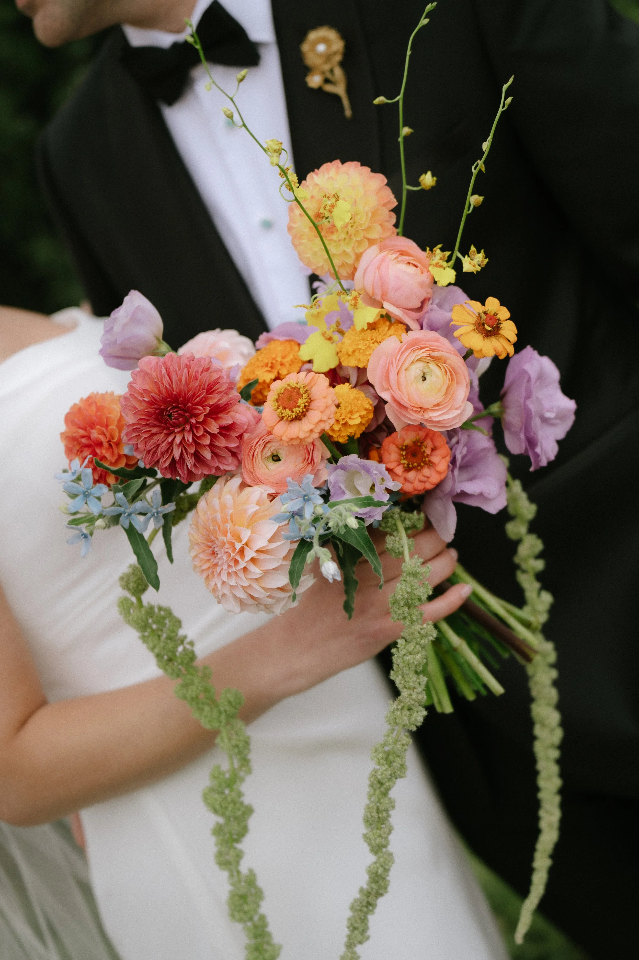 A person in a wedding dress holding a colorful bouquet of flowers, with a man in a tuxedo standing close by.