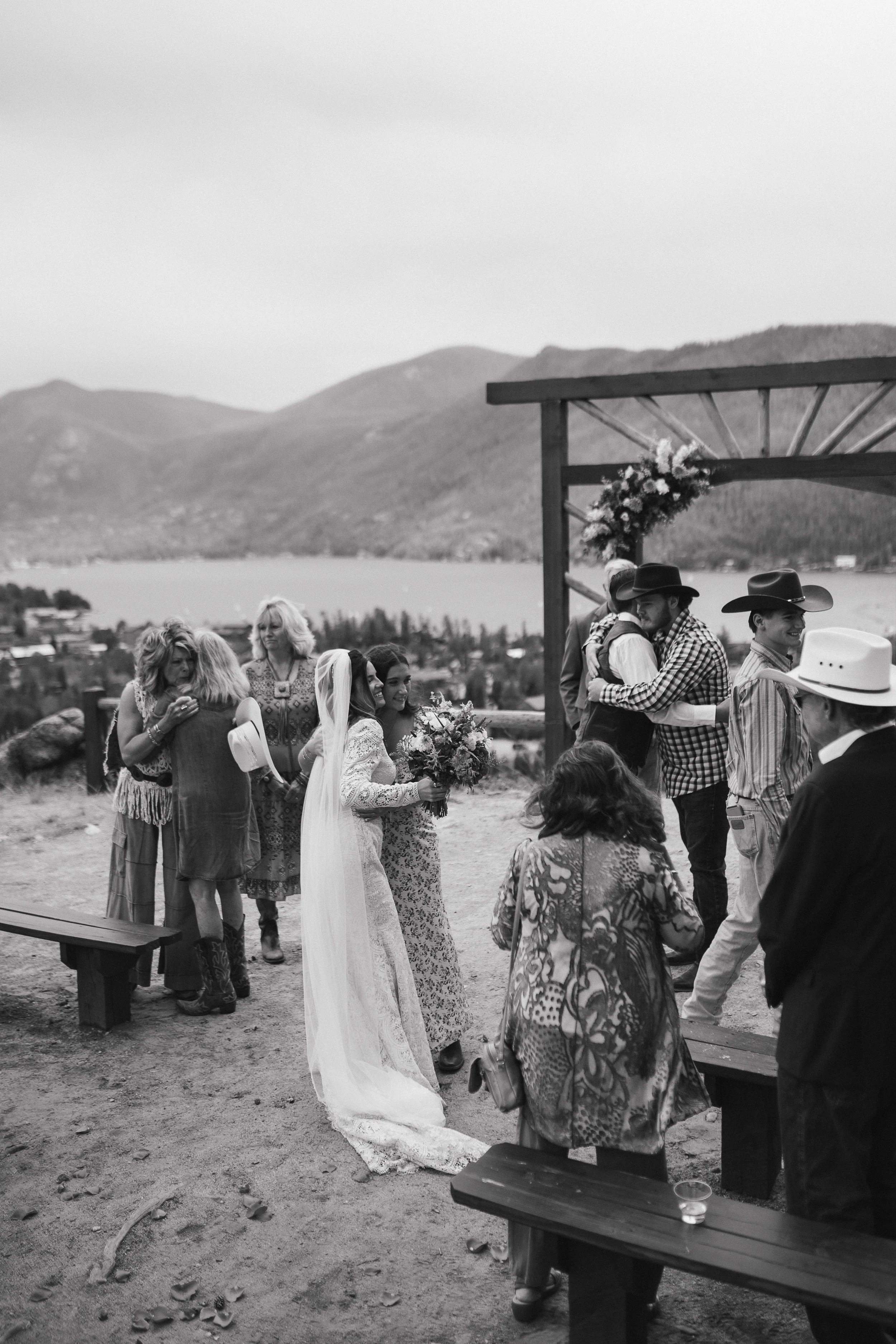 A black and white photograph of a wedding ceremony outdoors with a mountainous background. The bride is holding a bouquet and wearing a long white lace dress with a veil. Several guests are gathered around, some hugging. There is a decorated wooden arch and trees in the distance.
