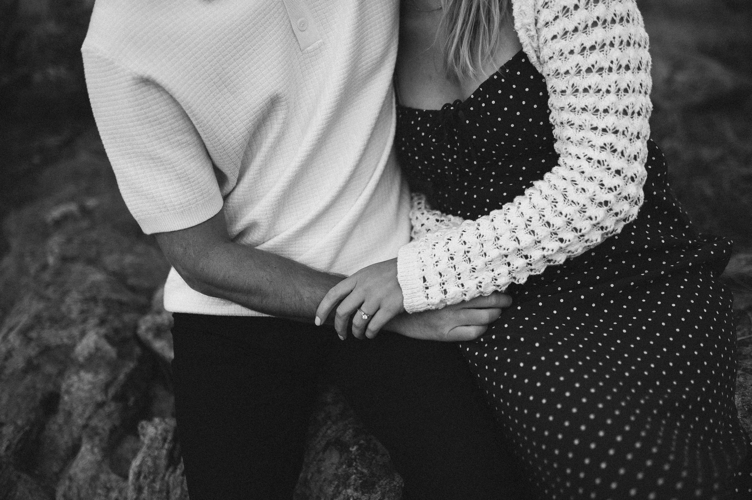 Close-up of a couple holding hands, showing only their torsos and arms. The person on the left wears a white short-sleeve shirt, and the person on the right wears a knitted sweater and a polka-dot dress.