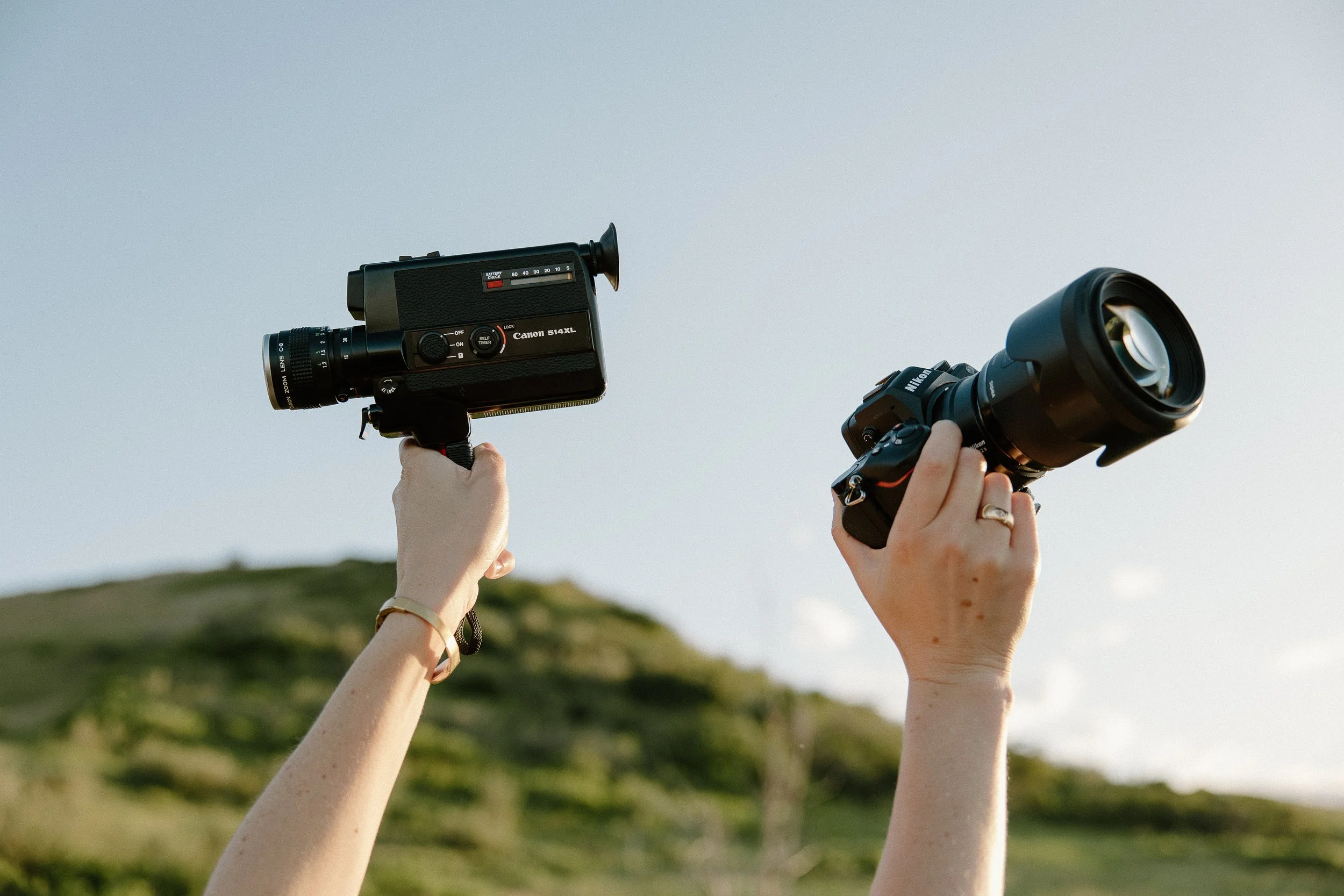 Two hands holding cameras, one with a vintage camera and the other with a DSLR camera, raised in the air outdoors with a grassy hill in the background.