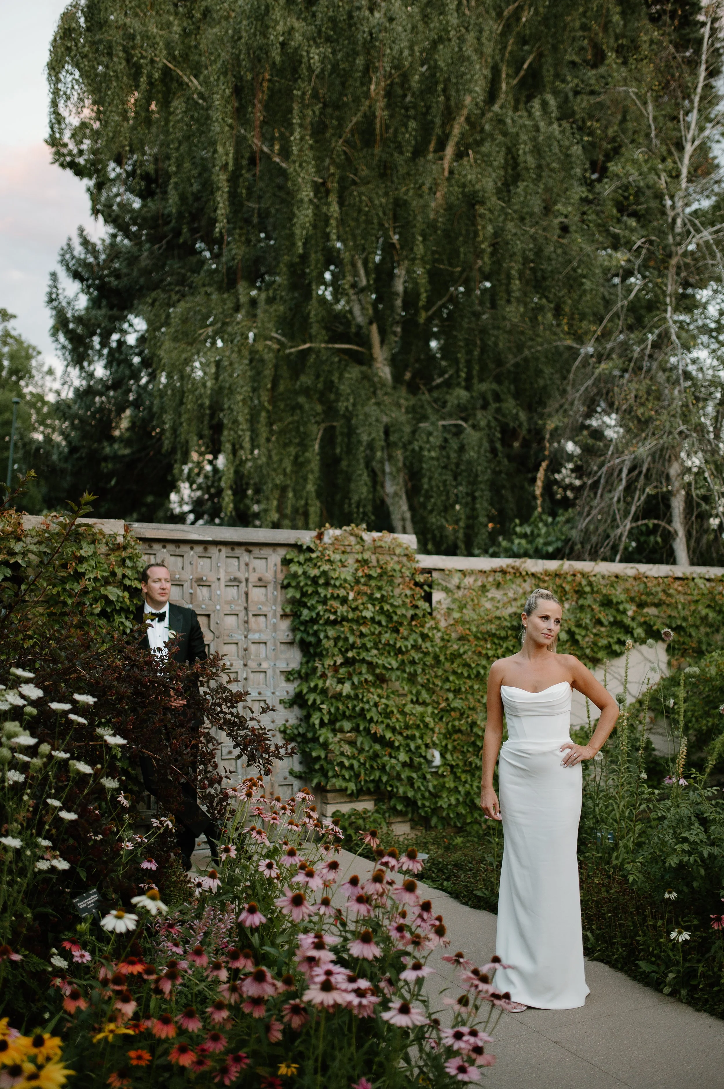 A woman in a white strapless wedding gown standing outdoors in a garden with colorful flowers and greenery, with a man in a tuxedo in the background.