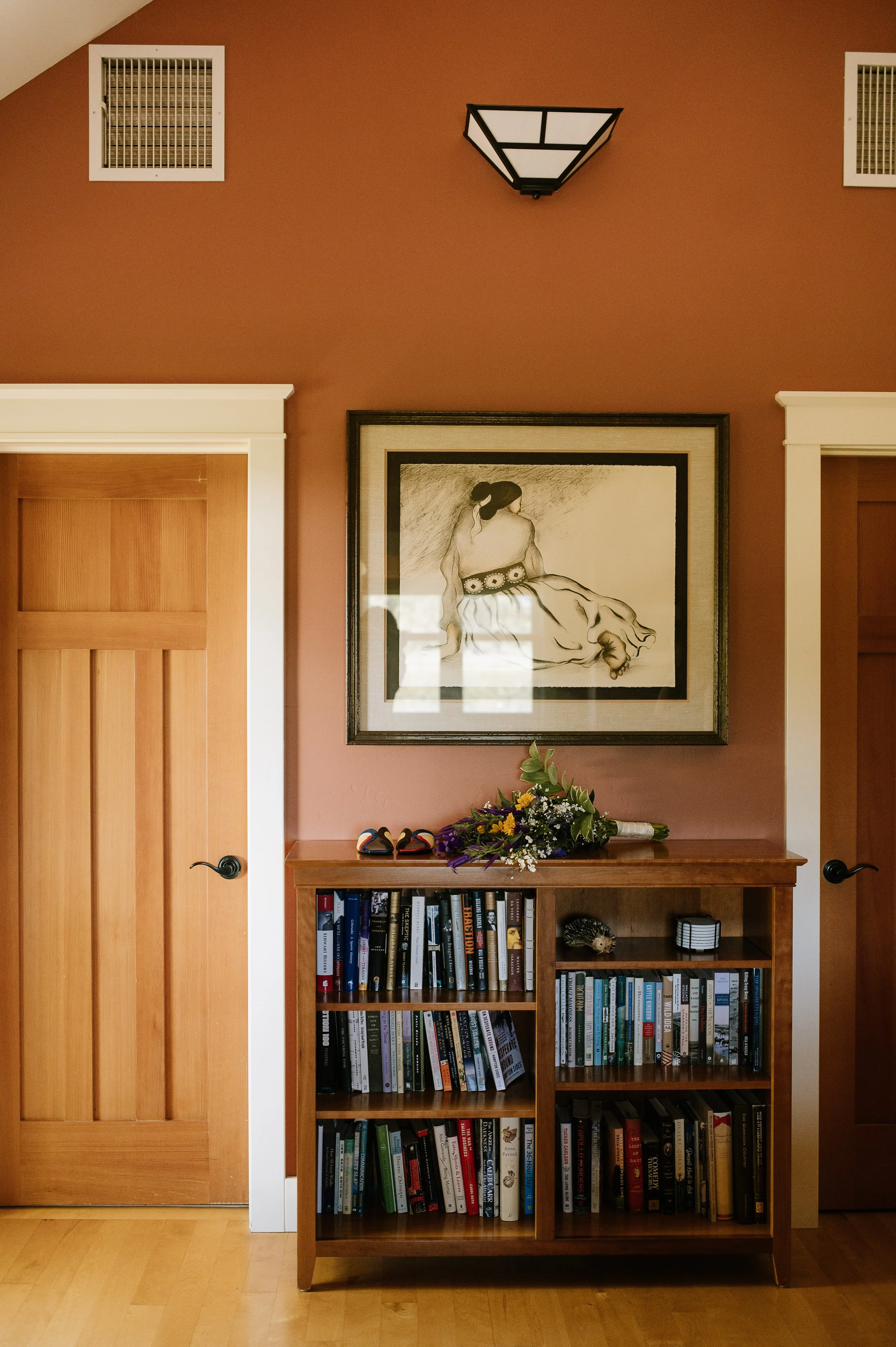 shoes and a bouquet on a bookcase full of books beneath of charcoal drawing hanging on a wall
