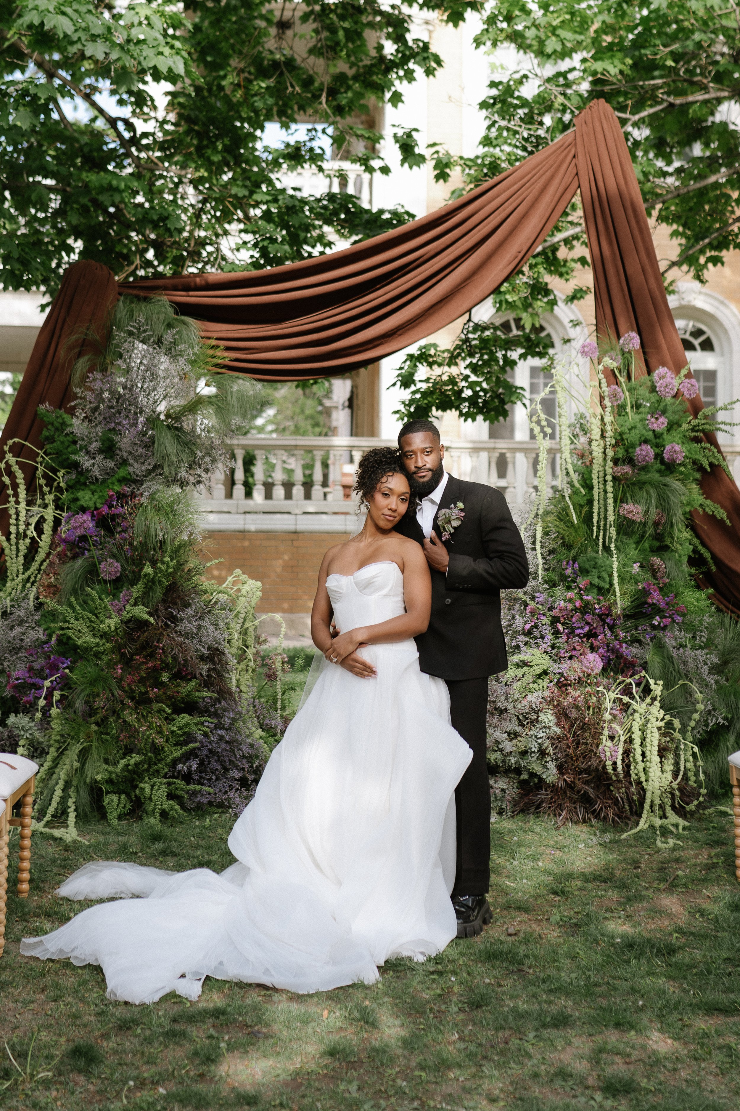 bride and groom posing for the camera with their floral ceremony arch in the background with purple flowers, greenery, and brown drapery