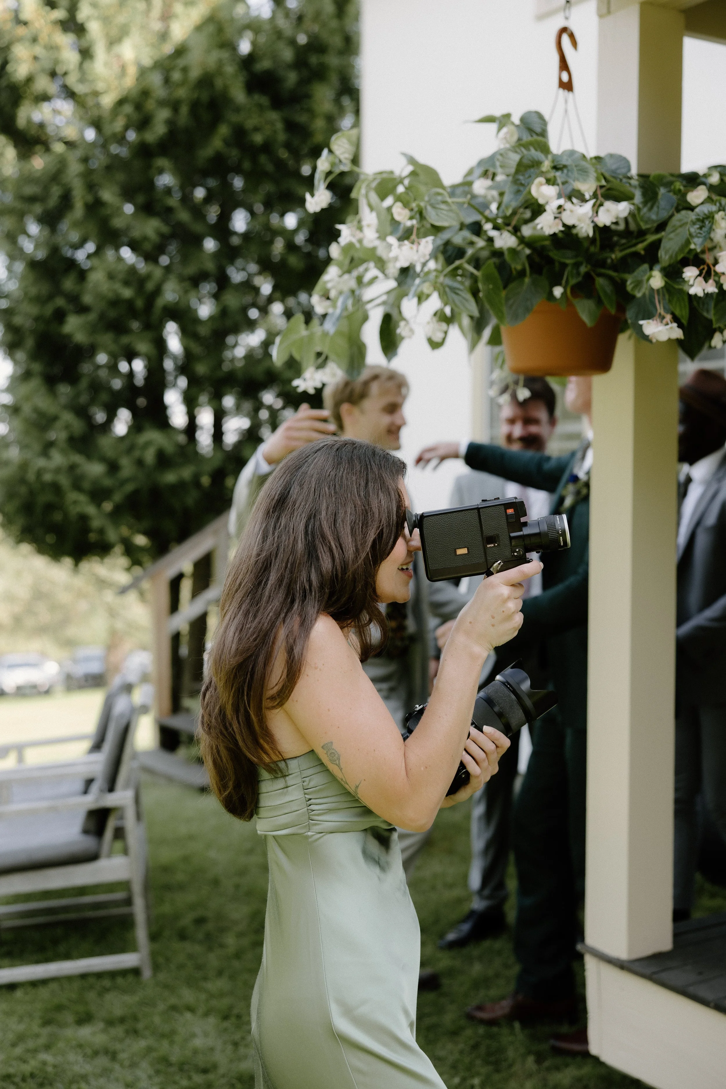 A woman with long brown hair taking a photo with a camera at an outdoor gathering during daytime. She is wearing a light green dress, and there are other people in the background smiling and talking near a porch with hanging flowers.