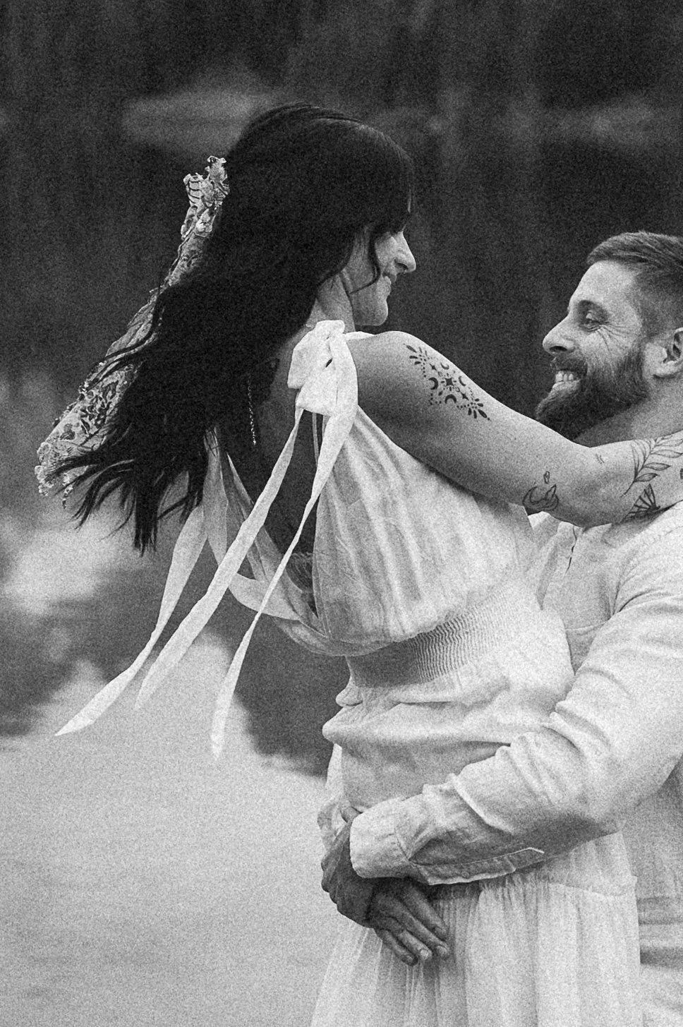 A black and white photo of a man lifting a woman, both smiling and looking at each other. The woman has tattoos and is wearing a dress with ribbons, while the man is dressed in a light-colored hoodie. They are outdoors, and the background is blurred.