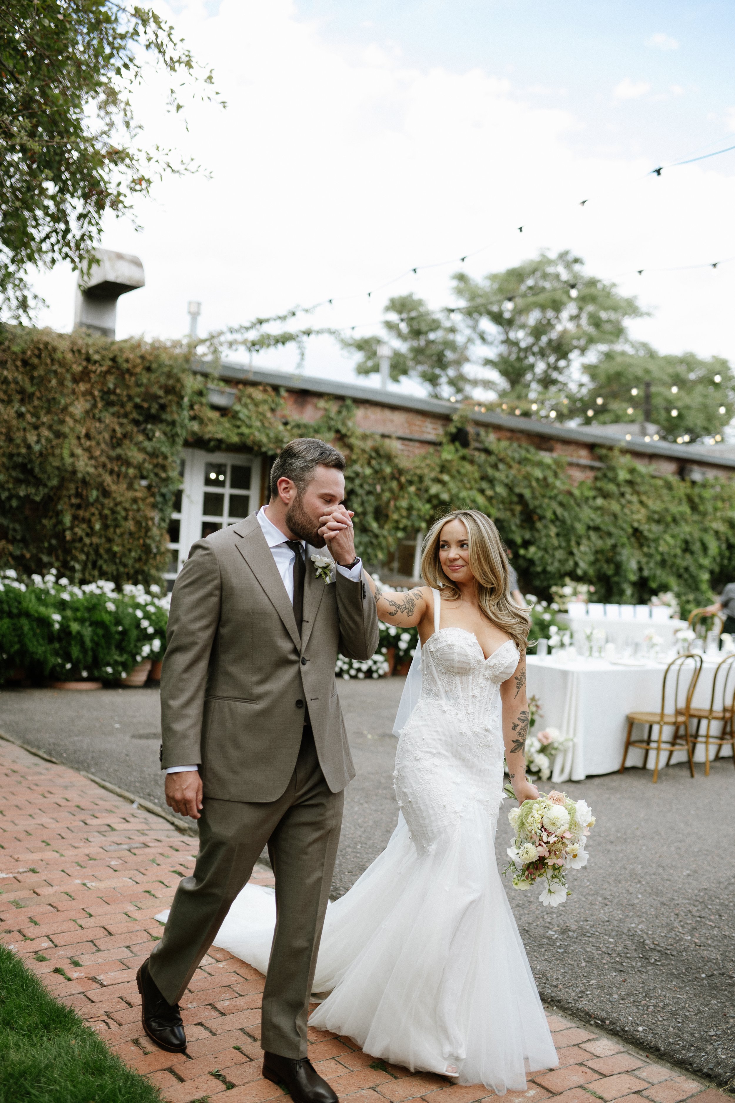 A bride in a white wedding gown holding a bouquet of white and pink flowers and a groom in a gray suit at an outdoor wedding reception. The groom appears to be wiping his eyes and the bride is smiling. There are string lights, a brick building covered in green vines, and white tables and chairs in the background.