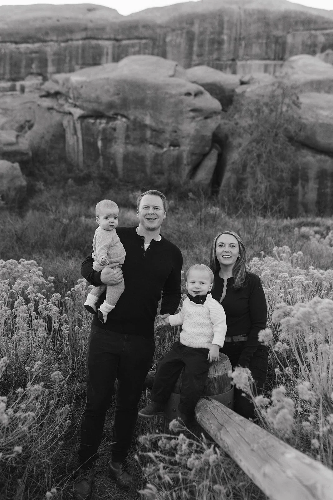 A family of four posing outdoors in front of a rocky, cliff-like landscape. The father is holding a young child, and the mother has a young child sitting on a log in front of her. All are smiling and surrounded by tall grass and flowers.