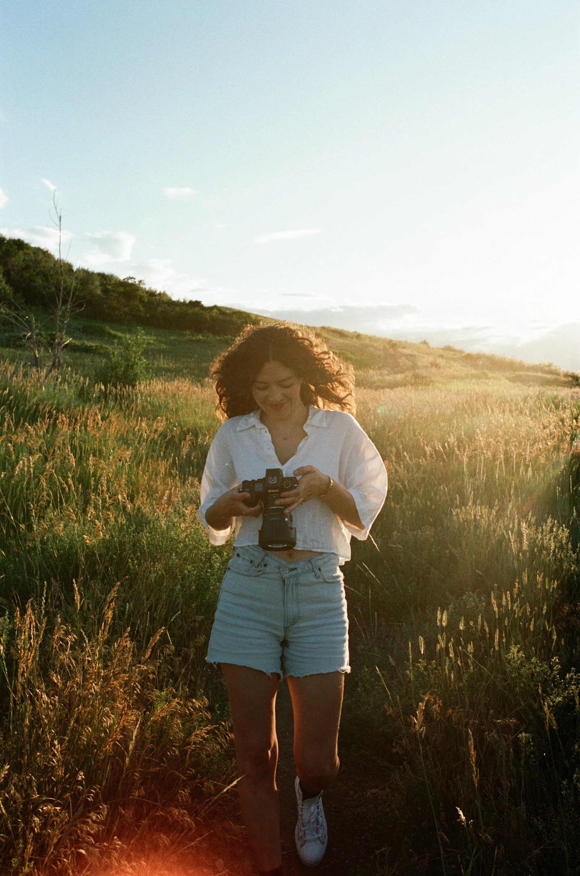 Young woman with curly hair smiling while holding a camera, walking through a field of tall grass at sunset.