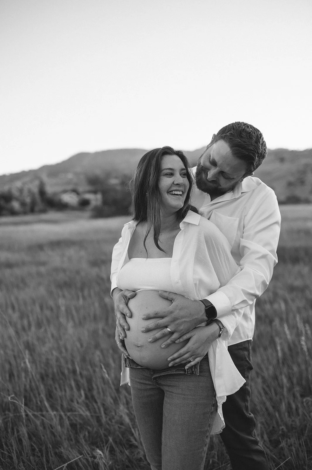 A happy couple, a pregnant woman and a man, standing outdoors in a field with mountains in the background, smiling and embracing each other.